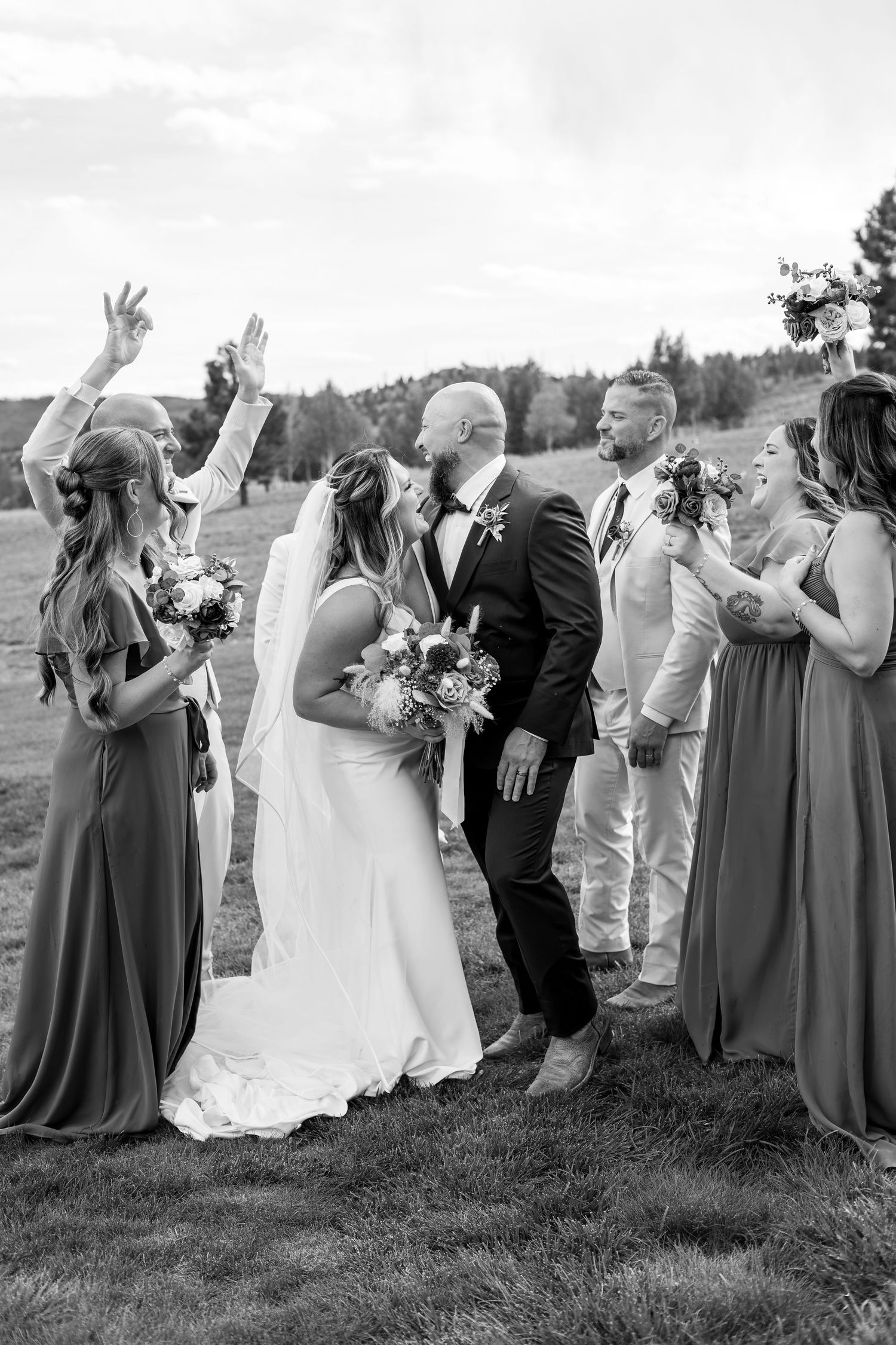 Black and white photo of bride and groom kissing surrounded by cheering wedding party outdoors.