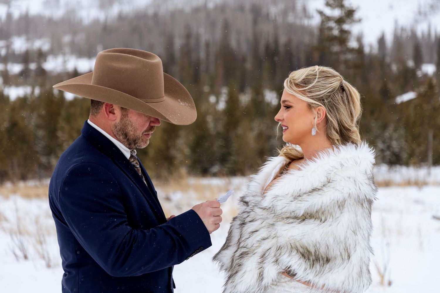 A couple shares a romantic moment in the snowy winter landscape wearing western attire and fur coats.