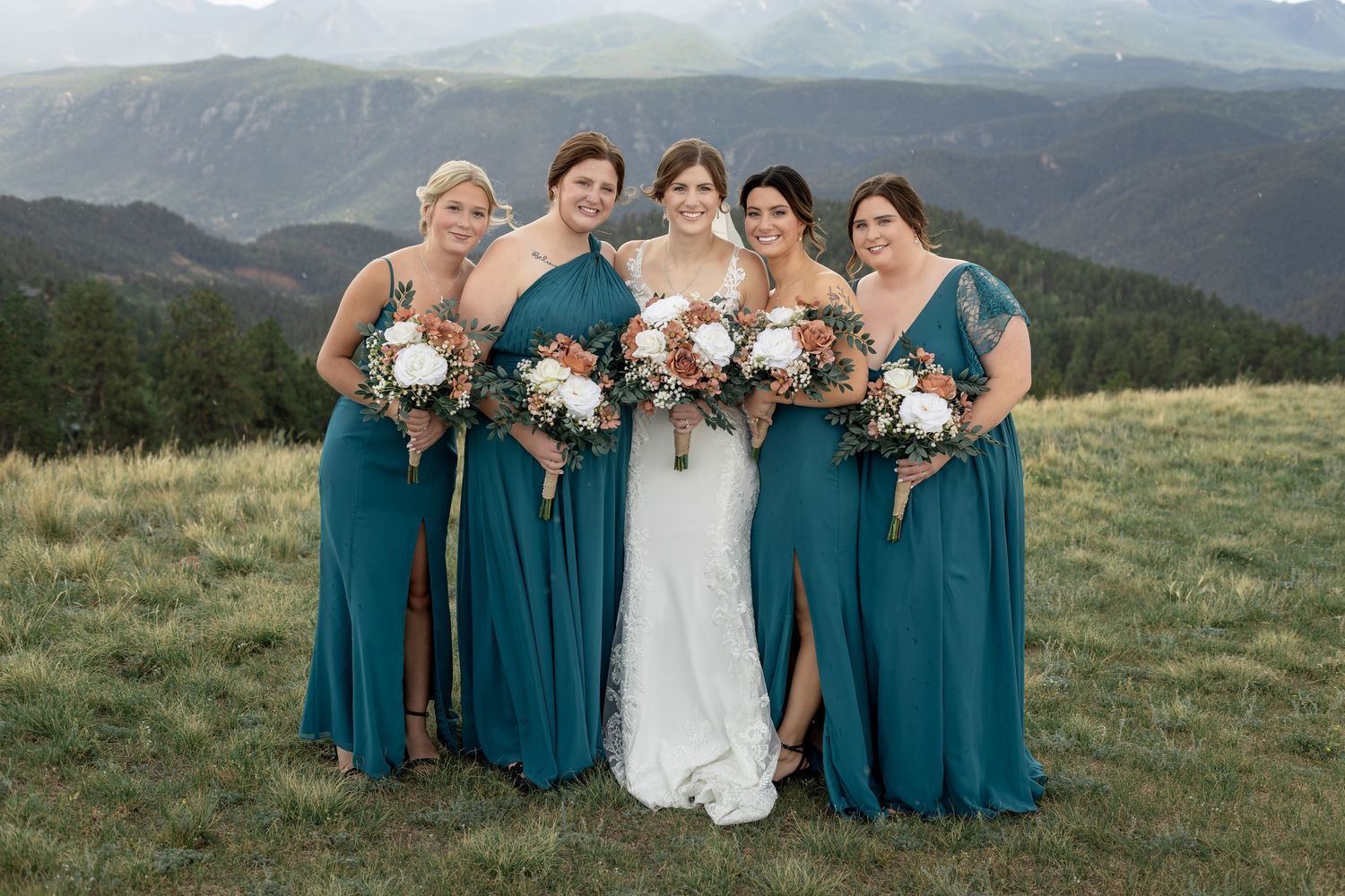 Bride in white gown with four bridesmaids in teal dresses holding bouquets on mountain meadow.