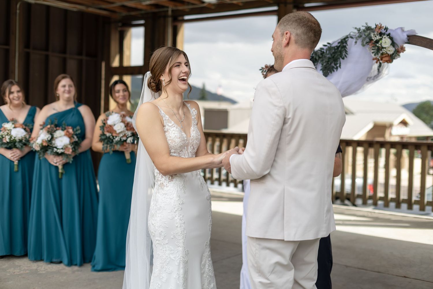 Bride laughing joyfully at groom during outdoor wedding ceremony with teal-dressed bridesmaids in background.