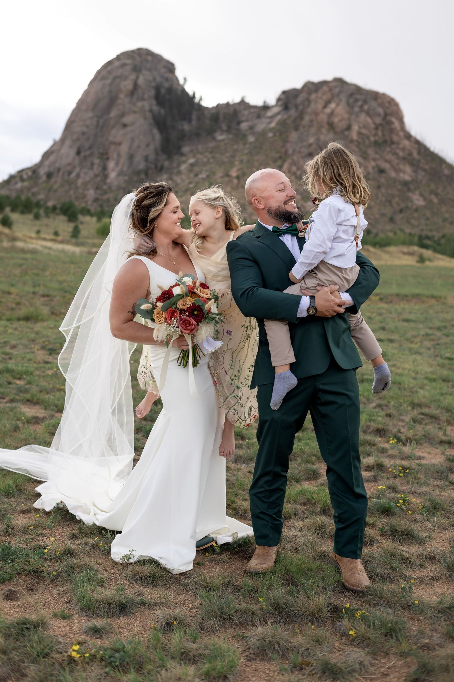 Bride and groom laughing with two children in a mountain meadow on their wedding day.