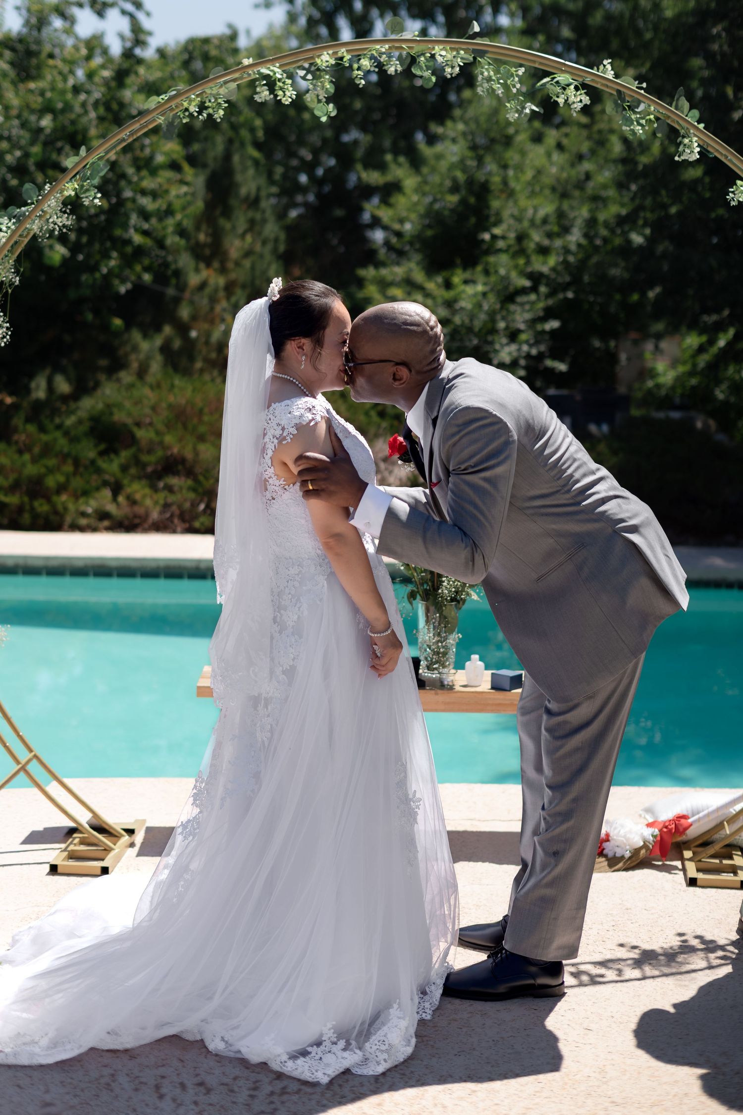 Interracial couple sharing first kiss at outdoor poolside wedding ceremony under floral arch.