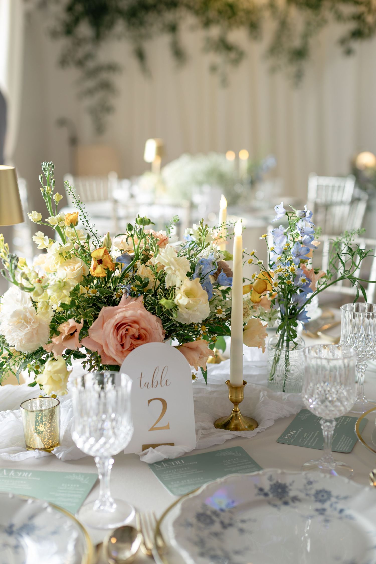 Elegant wedding table centerpiece with pastel pink, white, and blue flowers, gold candlesticks, and crystal glassware.