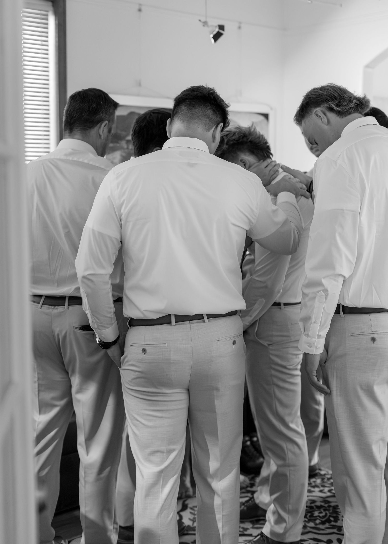 Black and white photo of groomsmen gathered together helping groom prepare before the wedding.