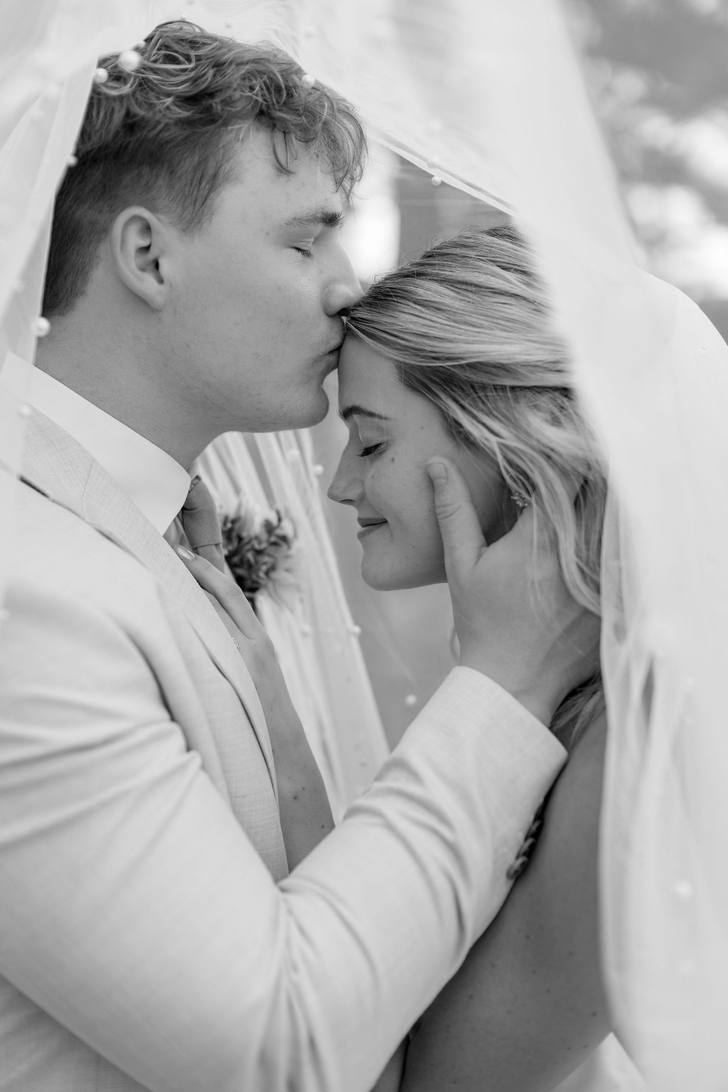 Black and white photo of groom kissing bride on forehead under wedding veil.