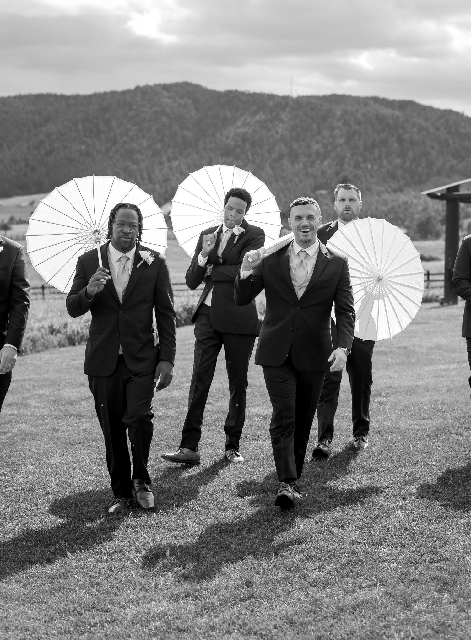 Groomsmen in black suits carrying white parasols walking across green field at outdoor wedding.