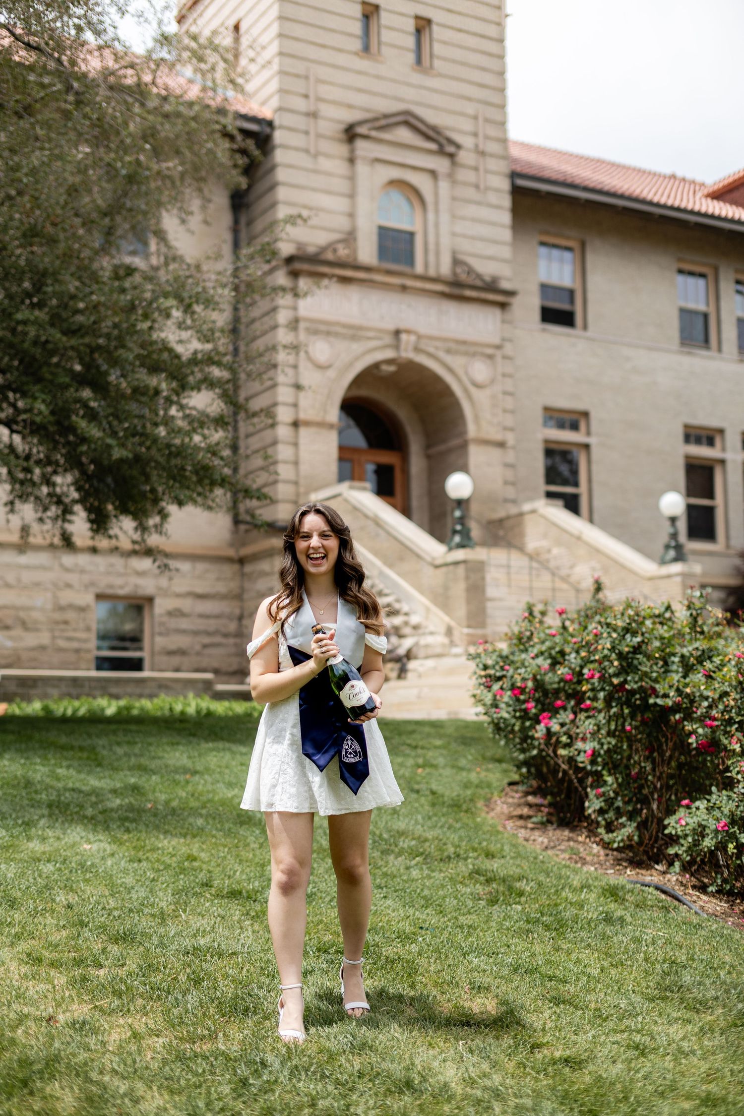 Smiling graduate in white dress holding champagne bottle with blue sash on university campus lawn.