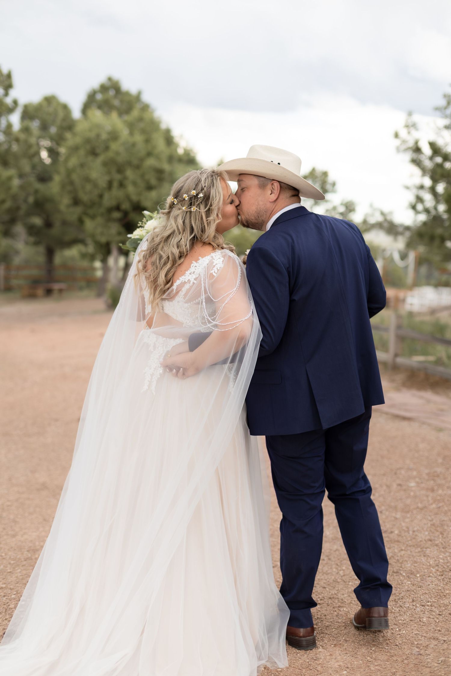 Bride and groom share a kiss outdoors, bride in white gown with veil, groom in navy suit and cowboy hat.