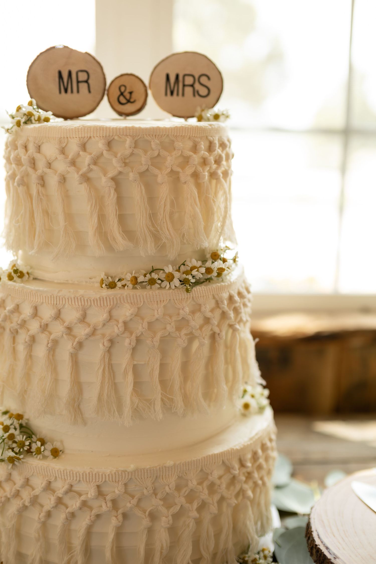 Three-tier white wedding cake with macrame fringe details and rustic MR & MRS wooden toppers.
