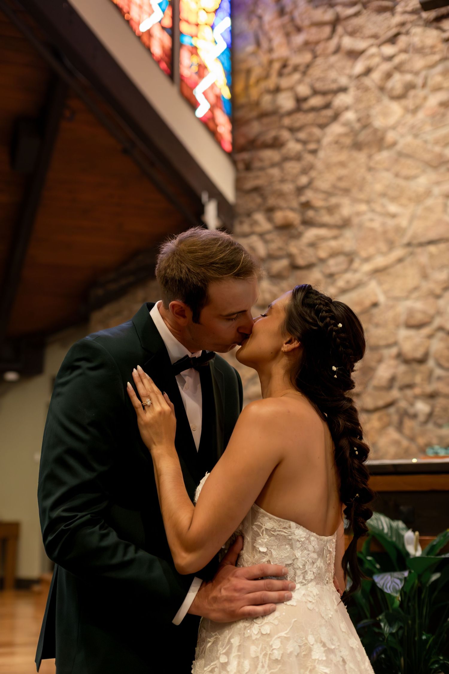 Bride and groom share their first kiss in a church with stained glass windows, bride in lace gown, groom in black tuxedo.