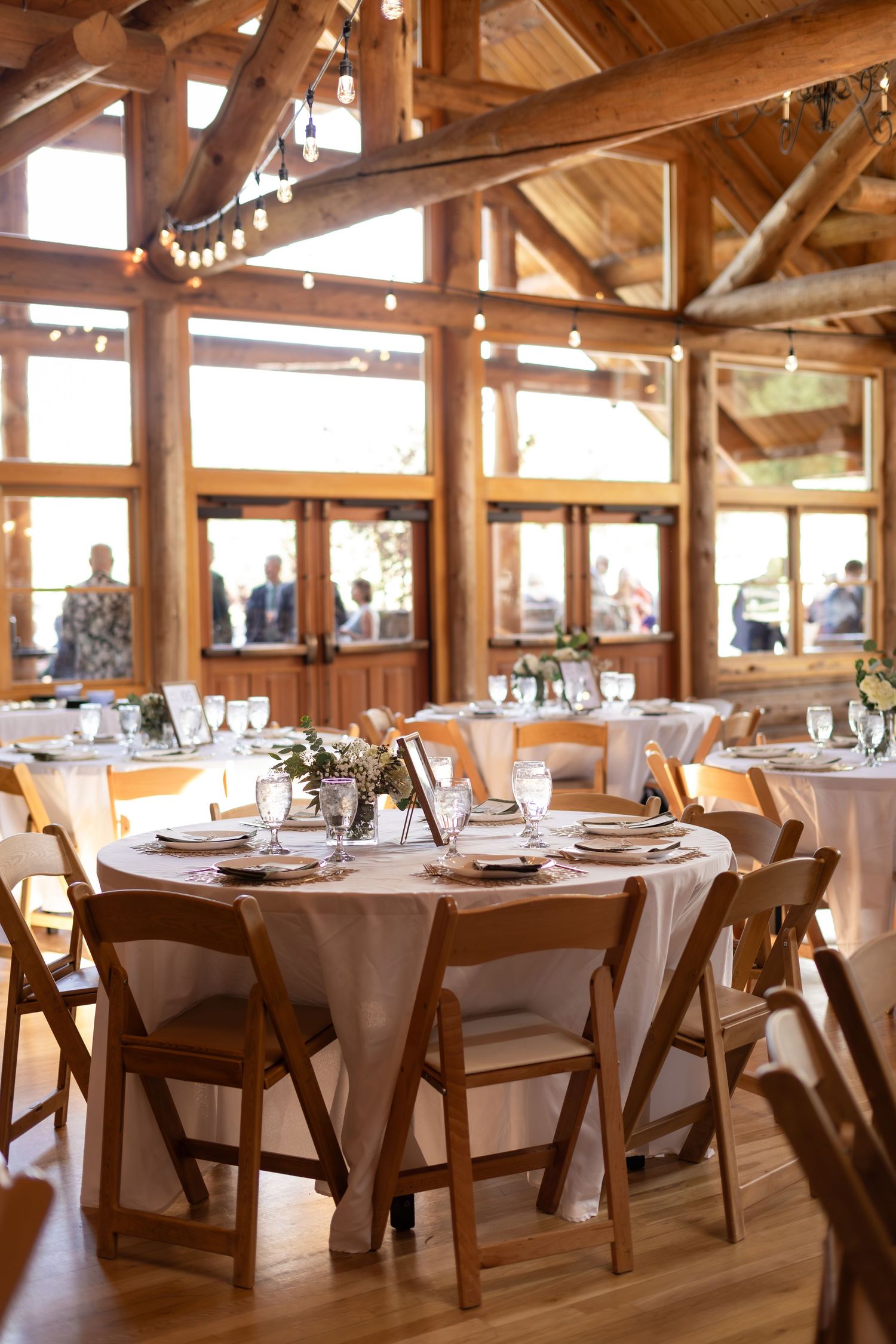 Rustic barn wedding reception venue with wooden beams, string lights, round tables with white linens, and folding chairs.
