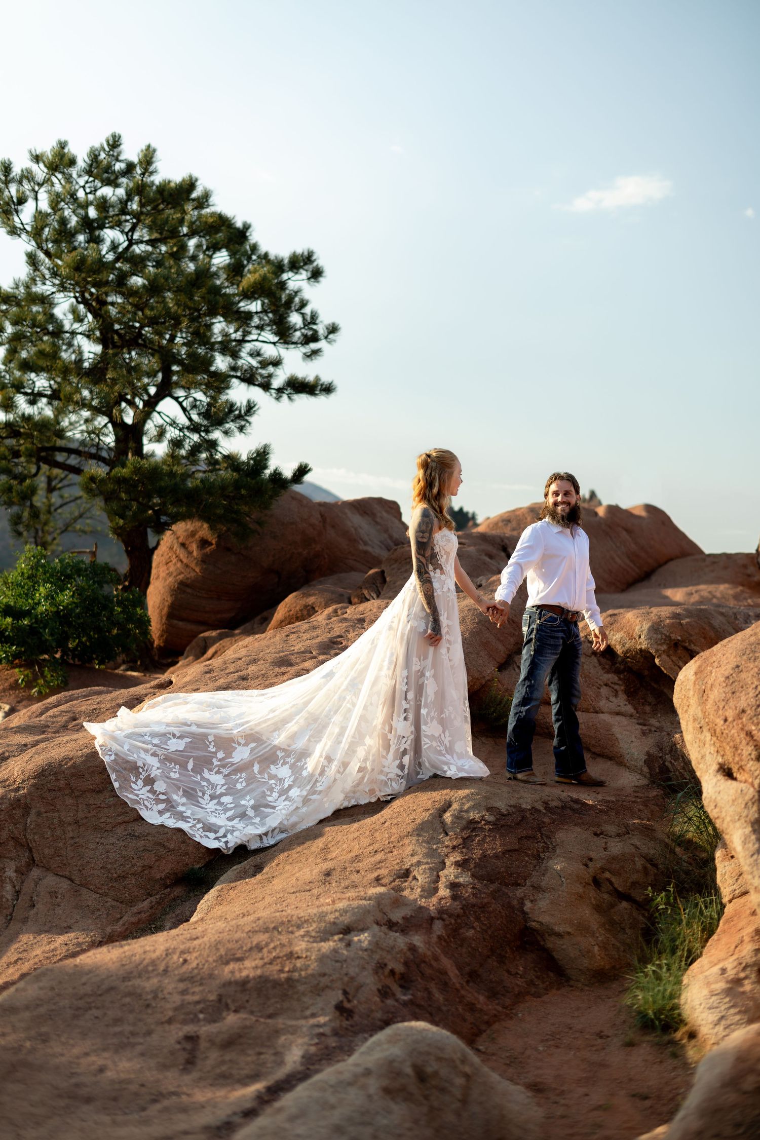 Bride in lace gown and groom in jeans hold hands on red rock formations with pine tree and blue sky backdrop.