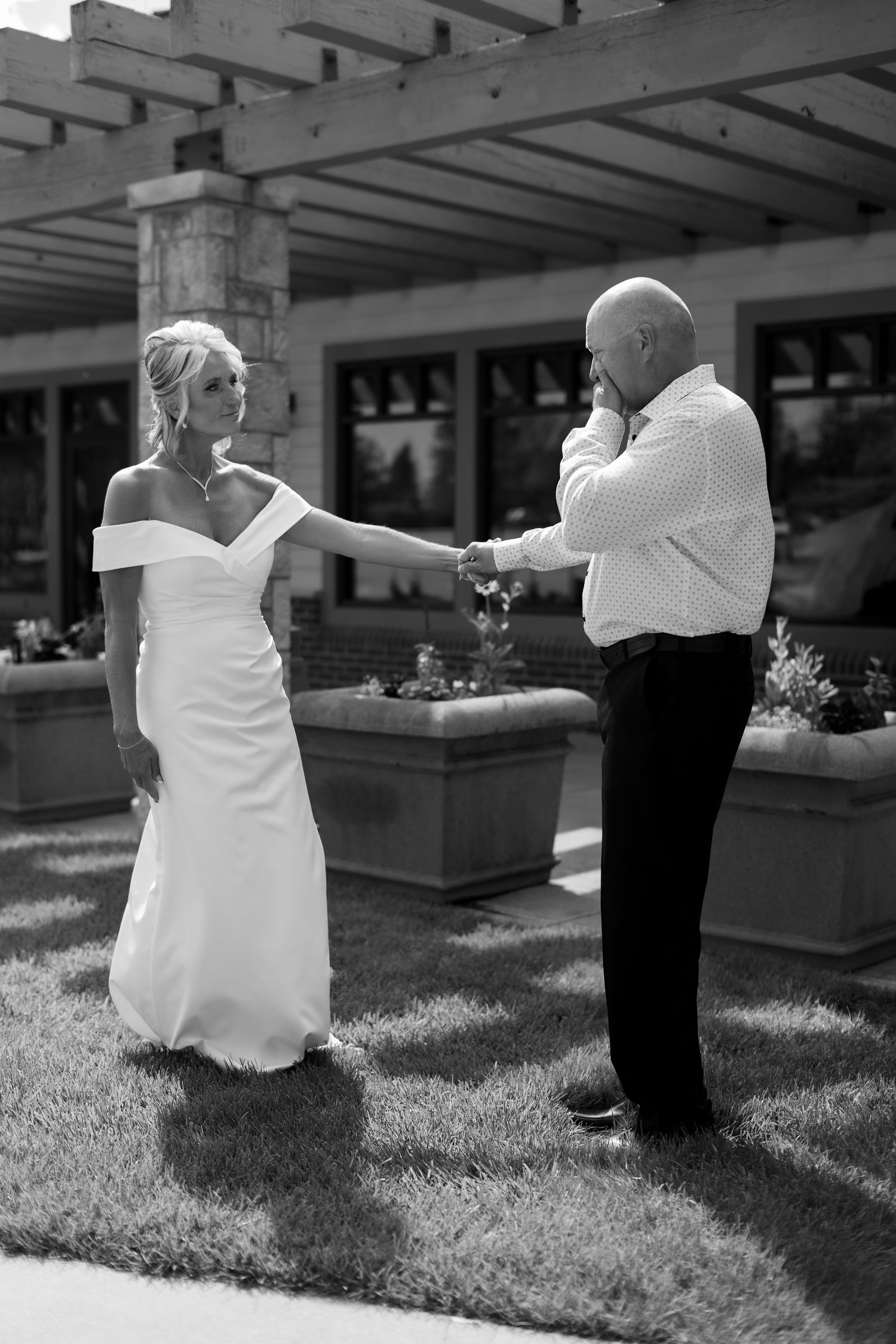 Black and white photo of a bride in white gown holding hands with emotional bald groom outdoors.