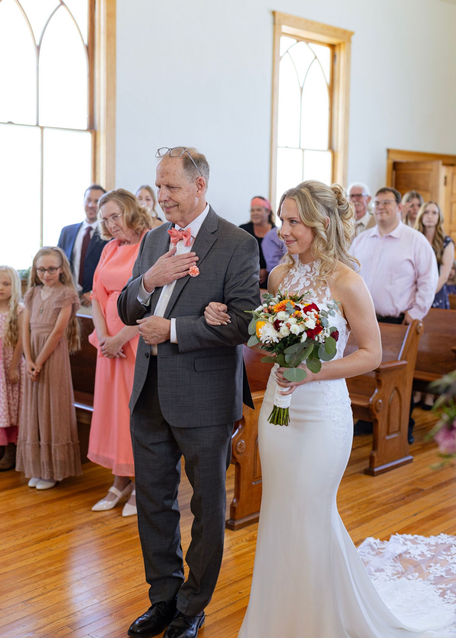 Bride in white lace halter gown walks down the aisle with her father holding a colorful bouquet.