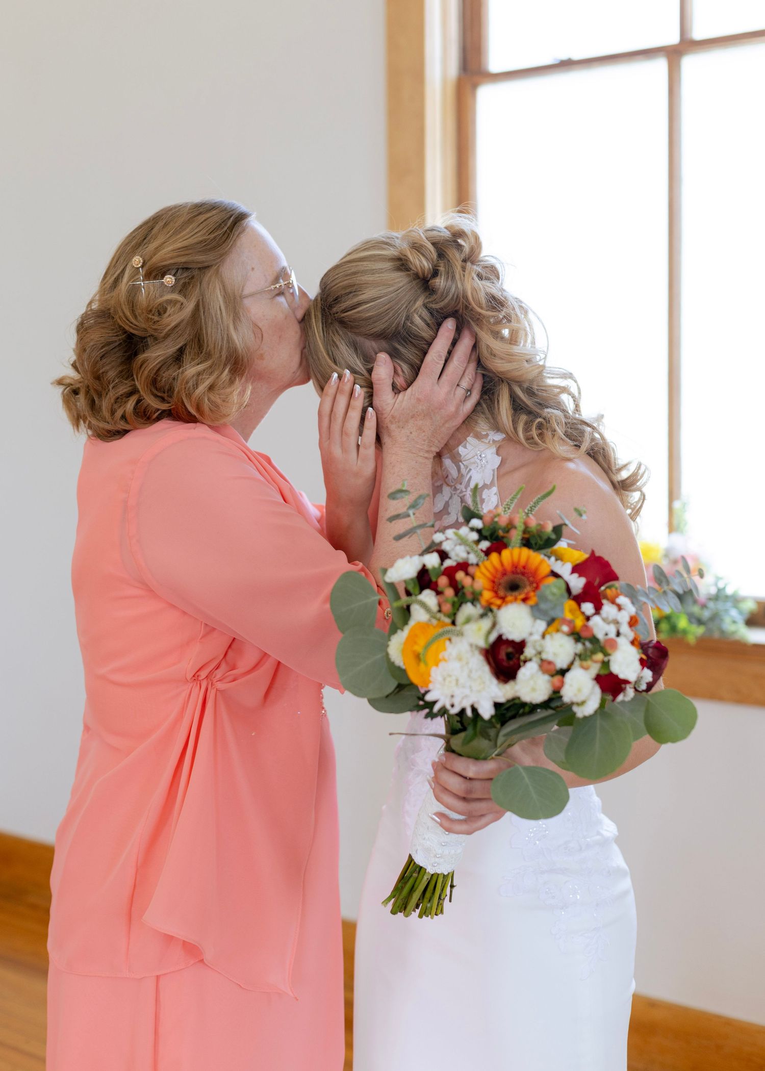 Emotional mother kissing bride on cheek while bride holds colorful fall wedding bouquet.