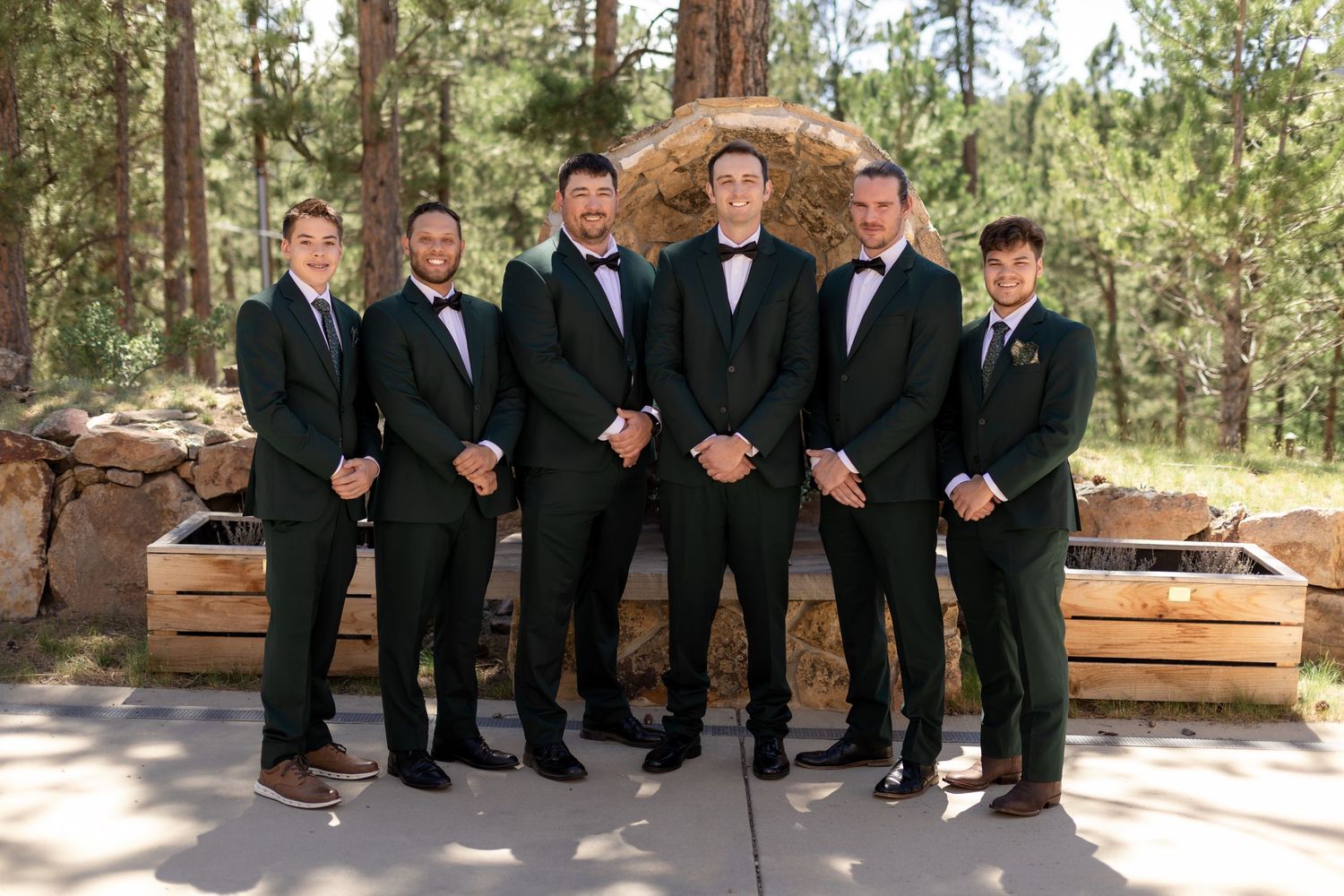 Six groomsmen in dark green tuxedos with black bow ties posing outdoors among pine trees at a mountain wedding venue.