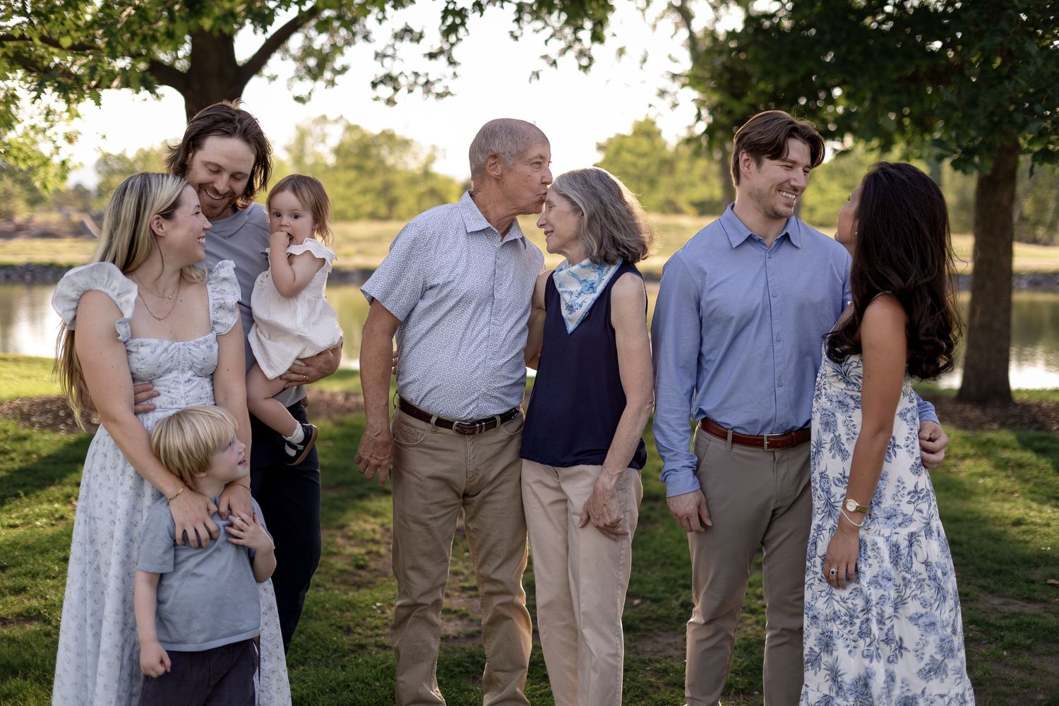 Multi-generational family gathering outdoors near a lake, smiling and interacting in casual attire on a sunny day.