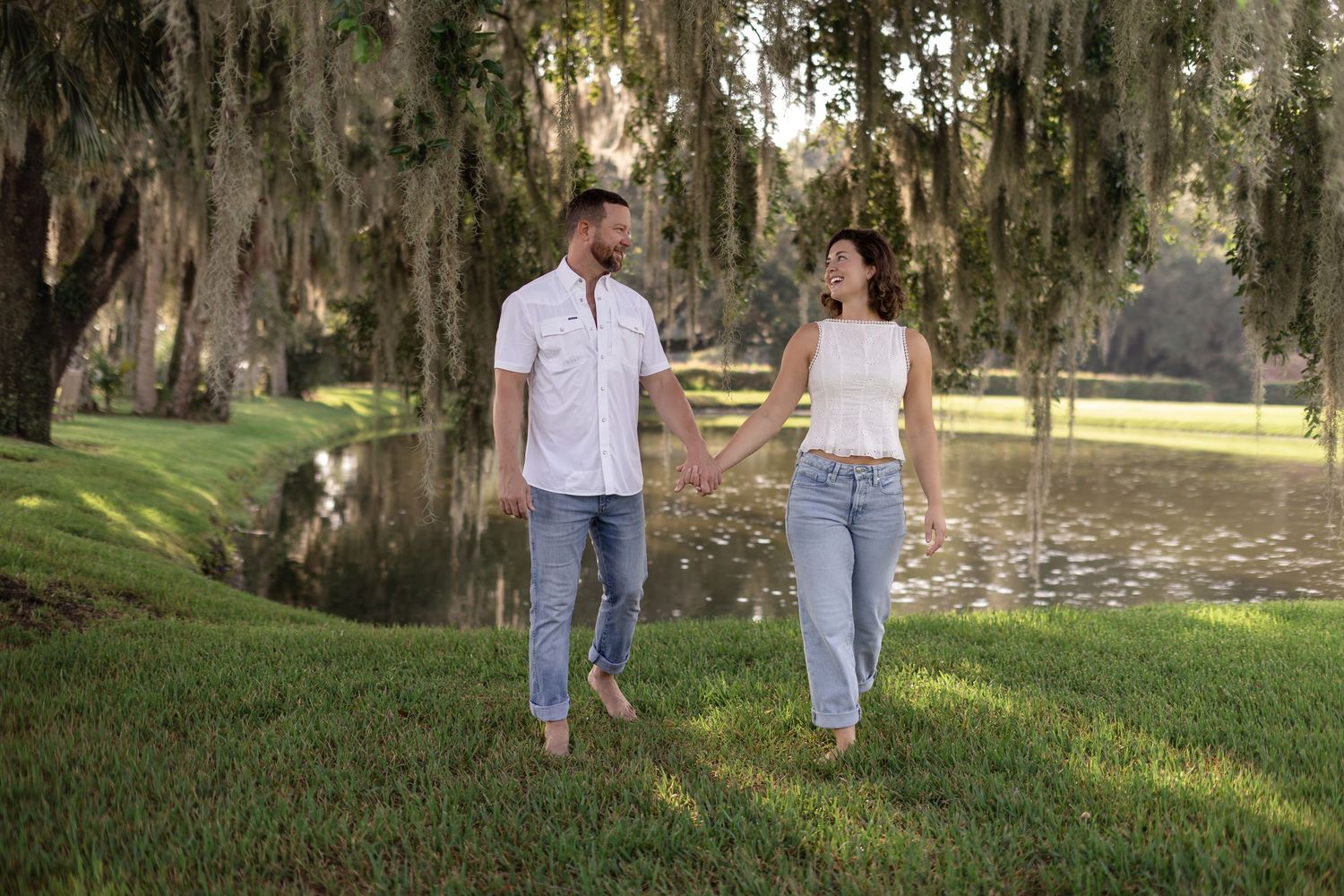 Couple holding hands walking on green grass near a pond under Spanish moss-draped trees in a park.