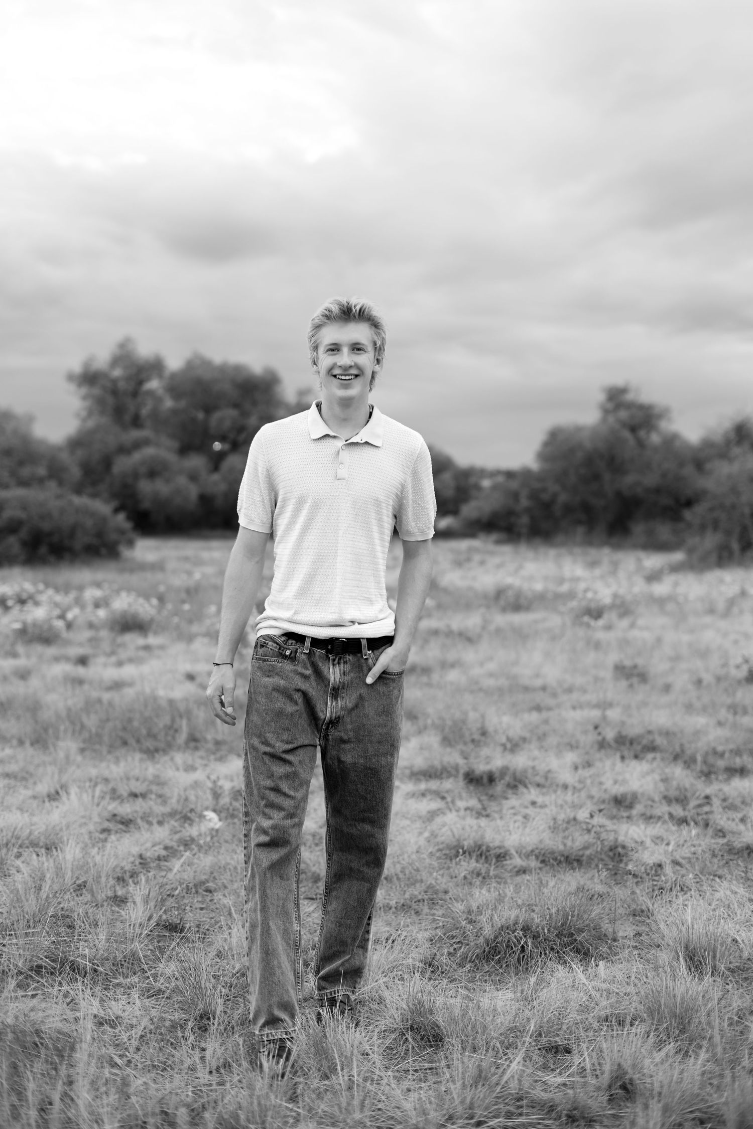Black and white photo of a smiling teenage boy standing in a grassy field with trees and cloudy sky behind him.