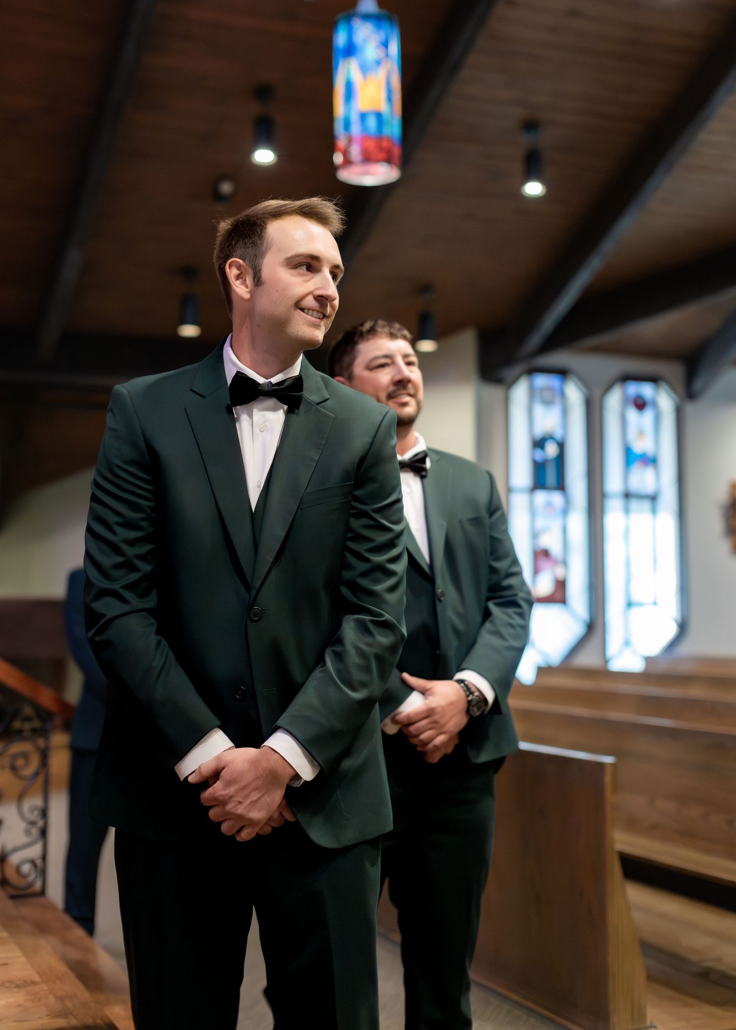 Two groomsmen in dark green tuxedos with bow ties stand at the altar of a church during a wedding ceremony.