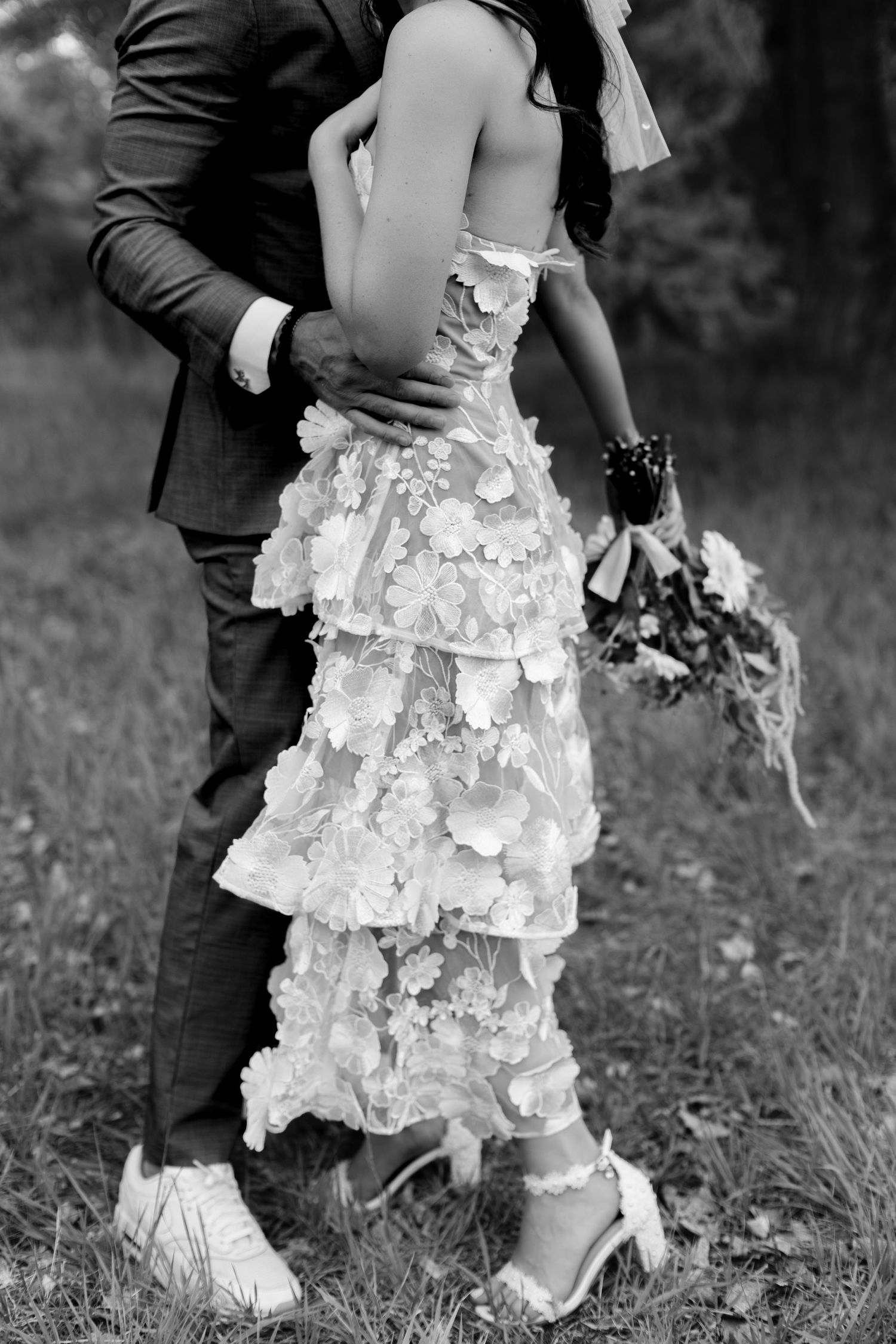 Black and white photo of bride in floral tiered gown embracing groom in suit outdoors.