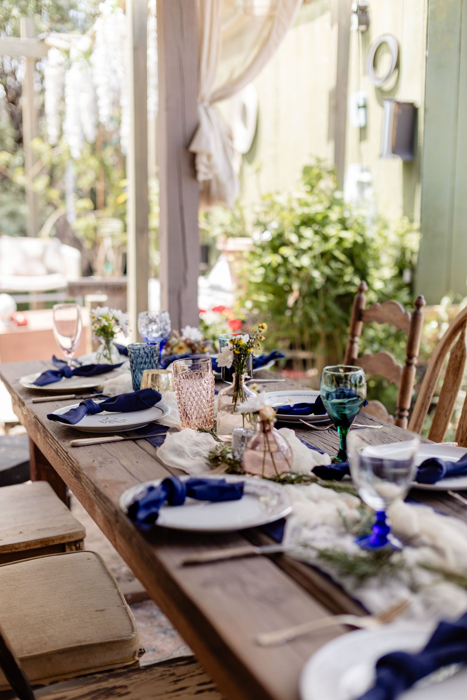 Rustic wooden dining table elegantly set with blue napkins, mixed glassware, and floral centerpieces in a sunlit garden setting.