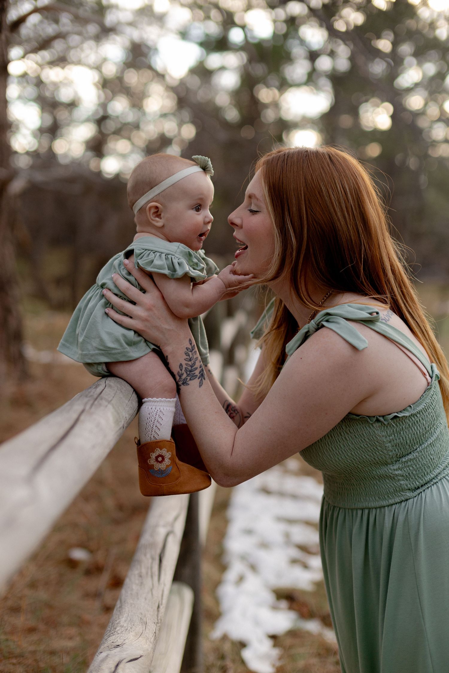 Red-haired mother in green dress holds smiling baby girl over wooden fence in a serene outdoor setting.