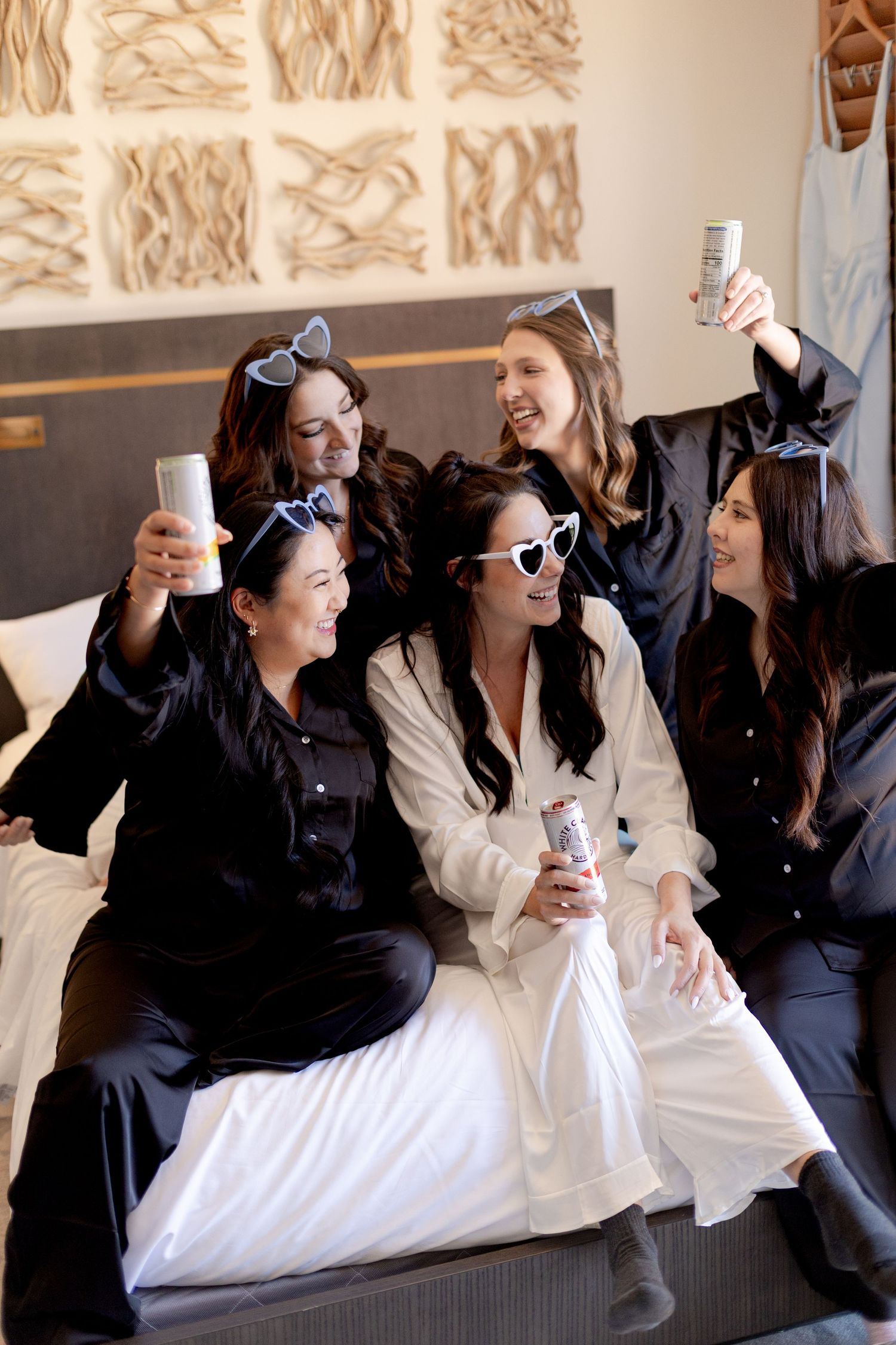 Bridesmaids in black satin pajamas celebrate with drinks on a hotel bed, laughing joyfully during bridal party getting ready.