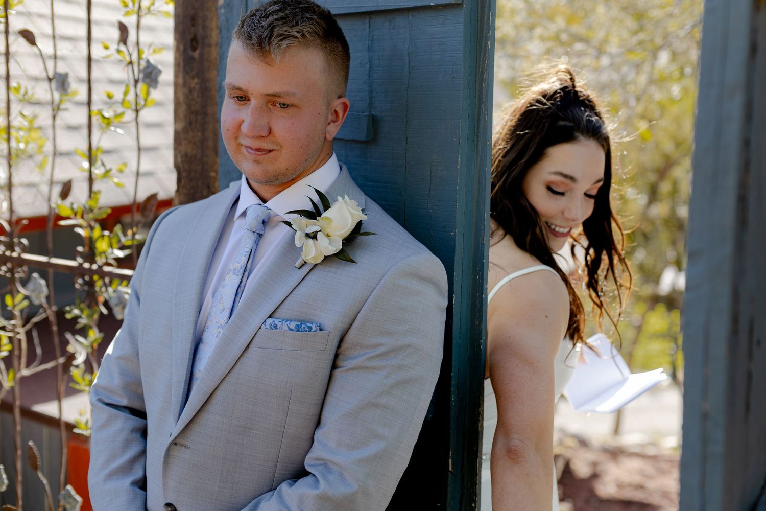 Groom in grey suit with boutonniere and bride in white dress leaning against blue wall before first look.