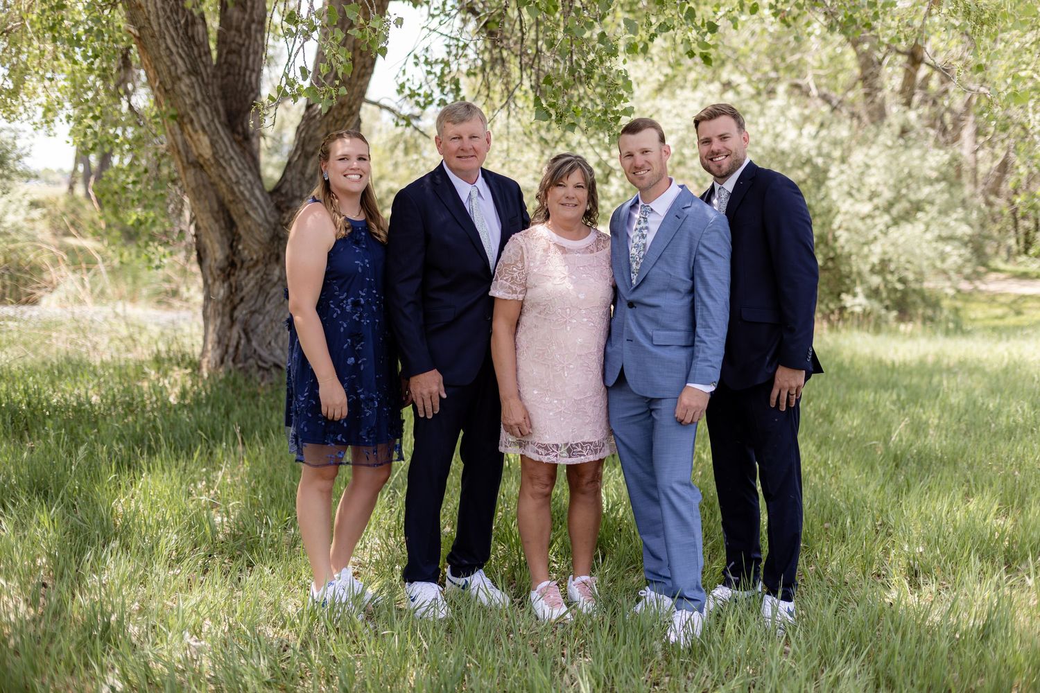 Family of five posing outdoors under a tree in a green meadow, dressed in formal attire for a special occasion.