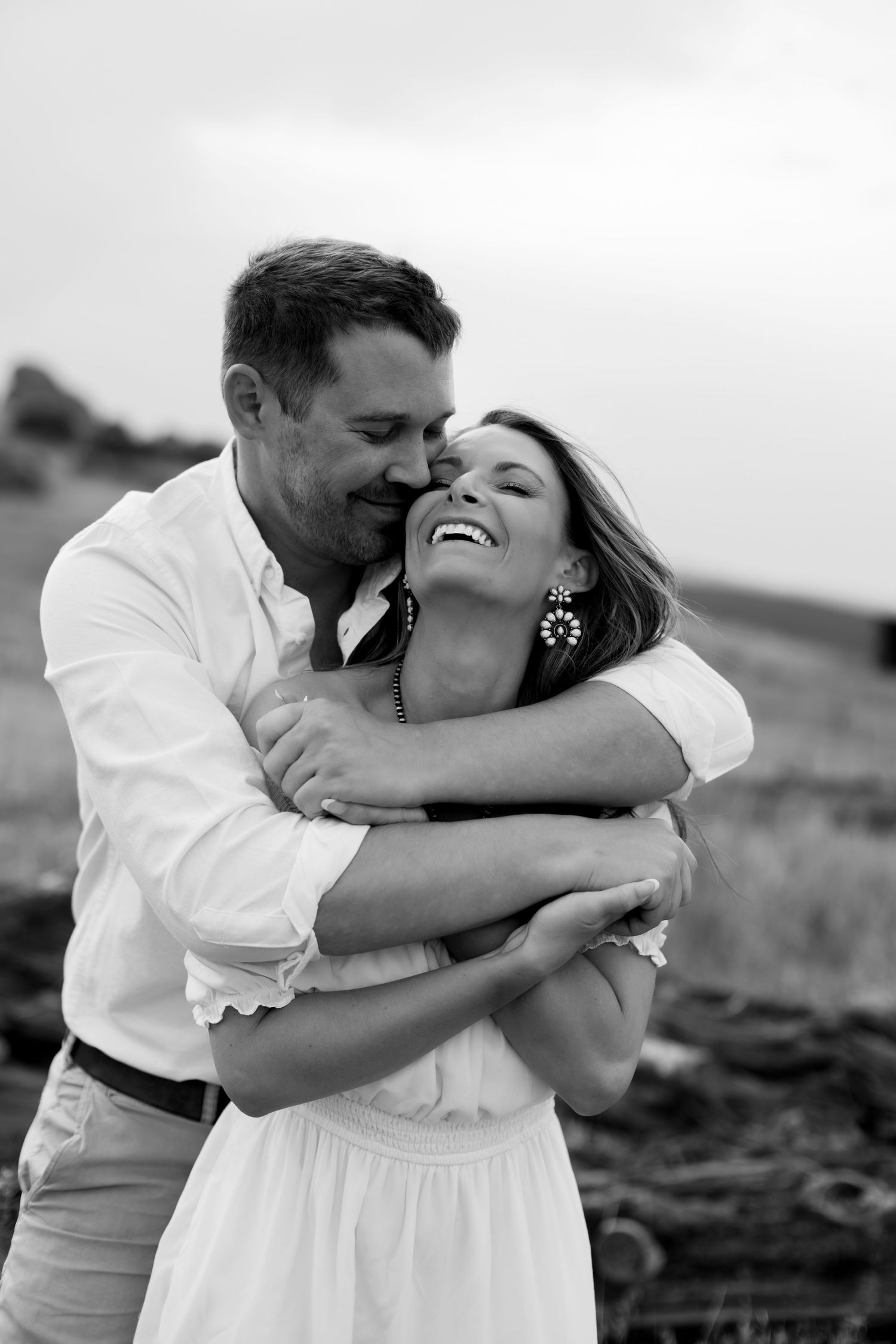 Black and white photo of a laughing woman being hugged from behind by her partner during an outdoor engagement session.