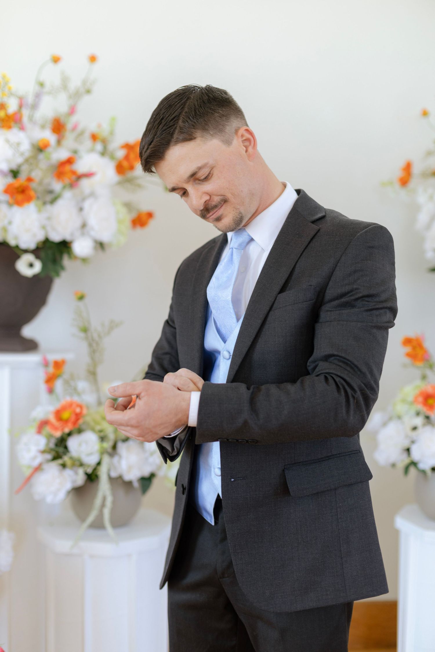 Dapper groom in dark suit and blue tie adjusting his cufflinks surrounded by orange and white floral arrangements.