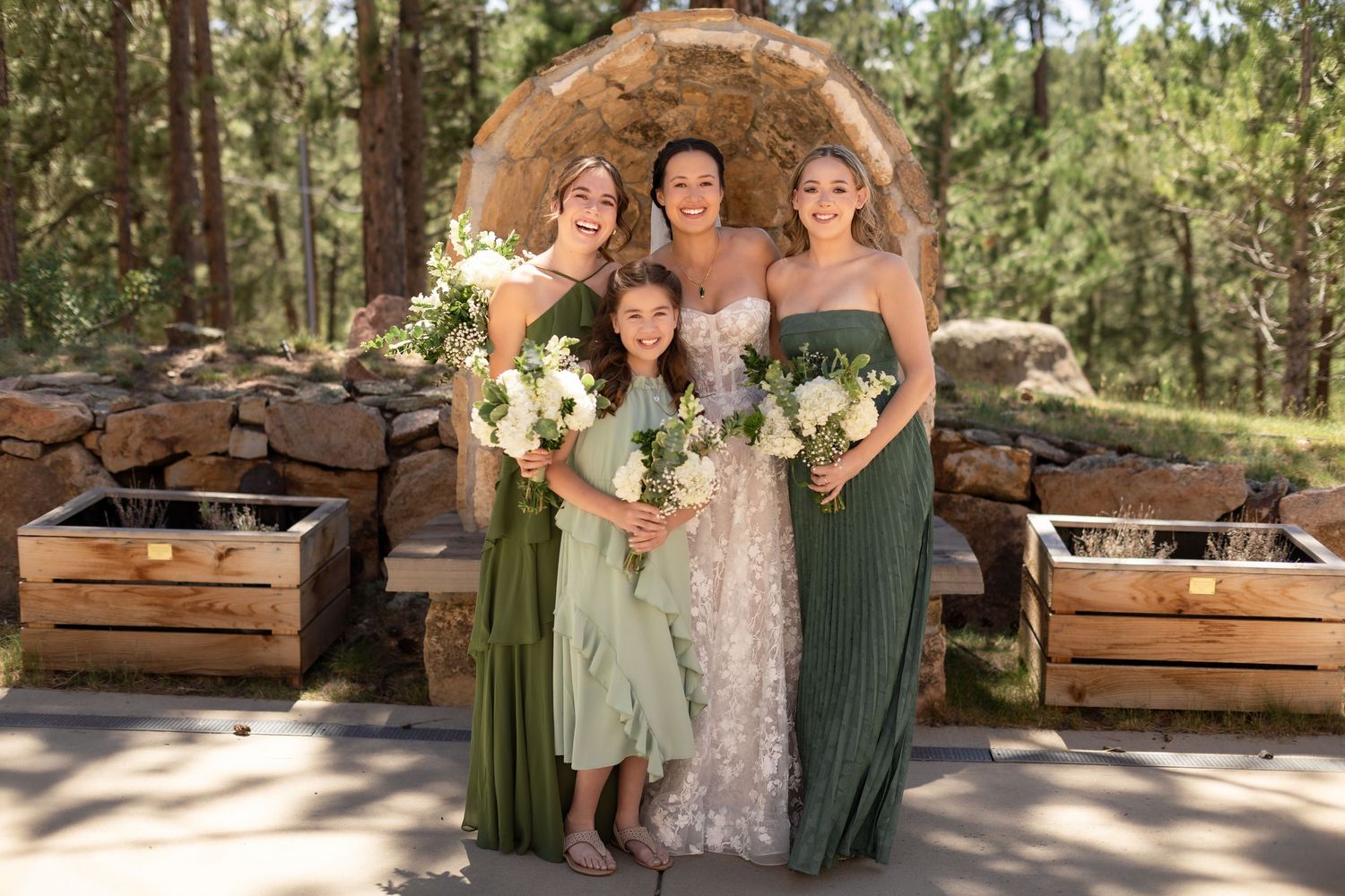 Bride in lace gown poses with two bridesmaids in sage green dresses and flower girl outdoors at rustic woodland wedding.