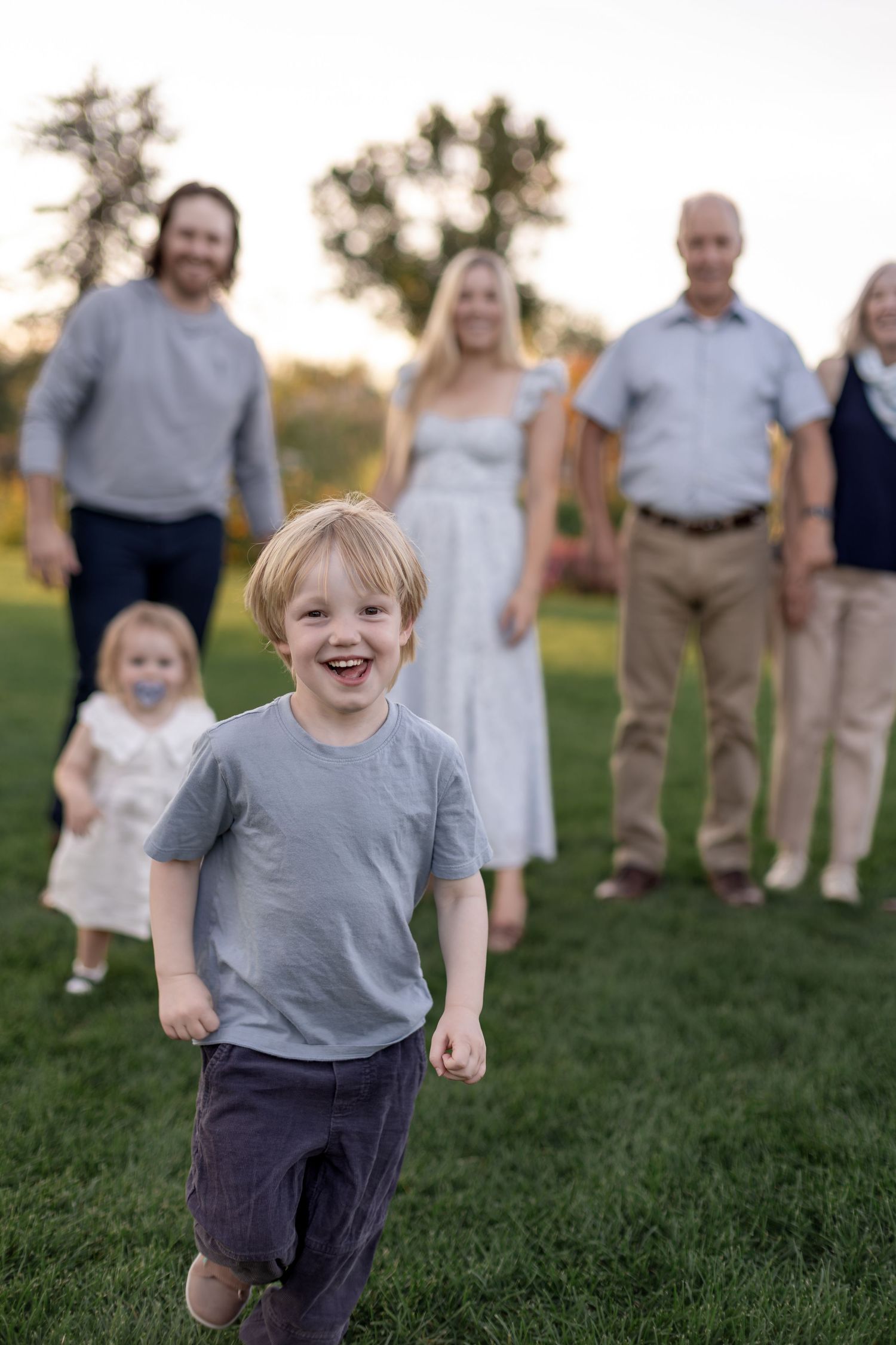 A joyful young boy runs toward the camera on green grass with his smiling family standing together in the background.
