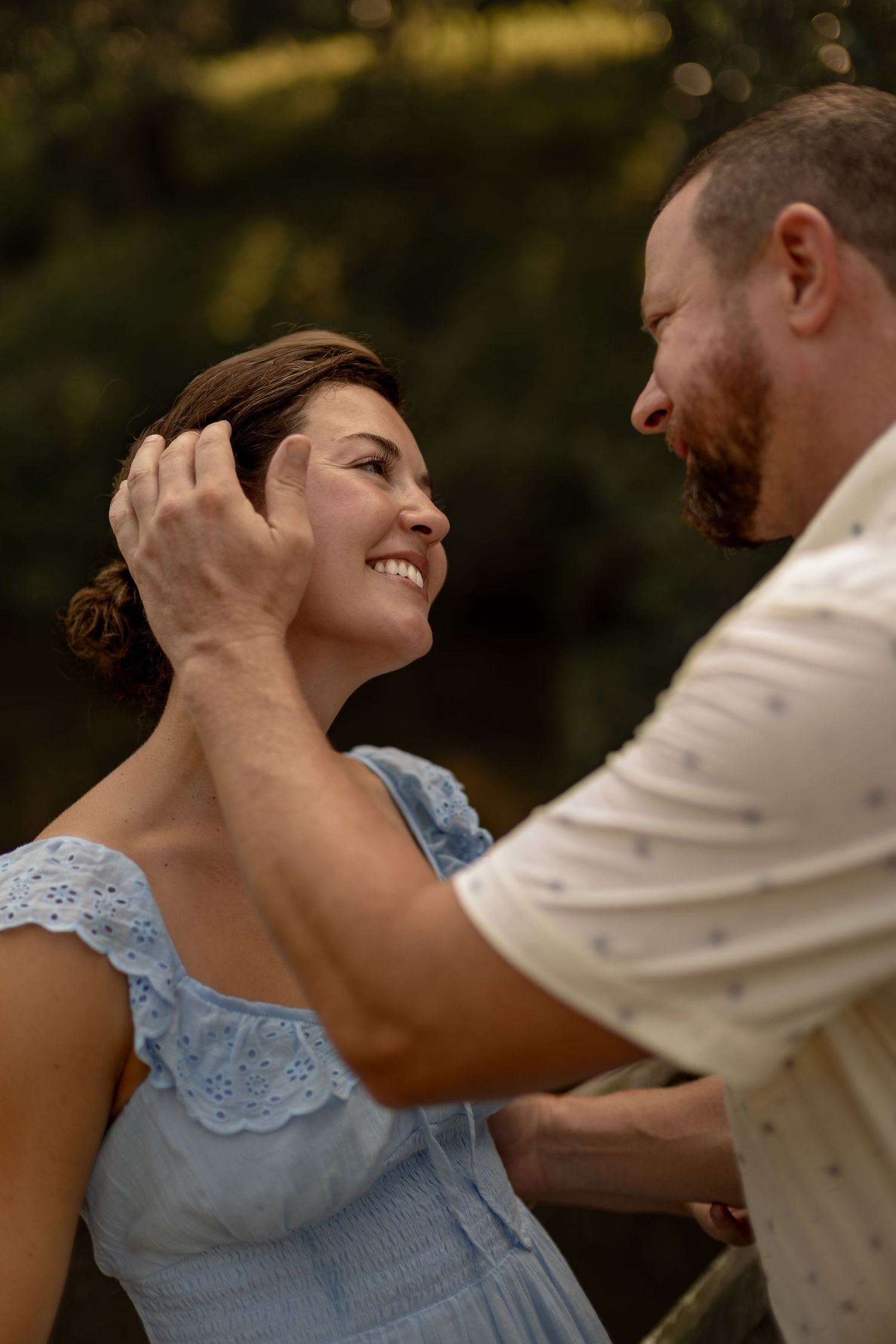 Smiling woman in blue dress gazes at man who gently brushes hair from her face outdoors.