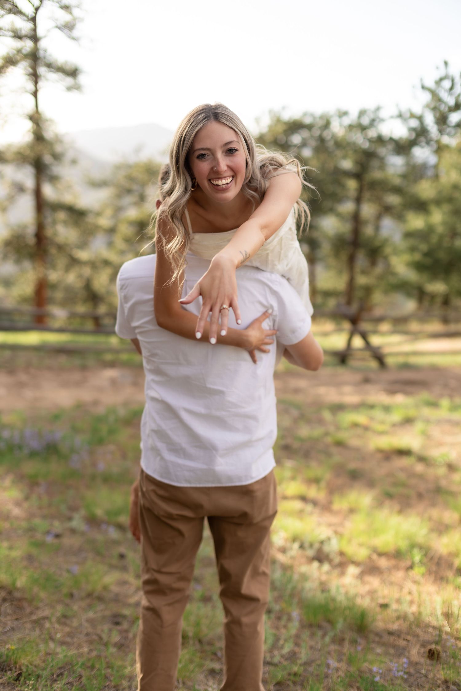 Smiling blonde woman showing engagement ring while being held by man in white shirt outdoors.