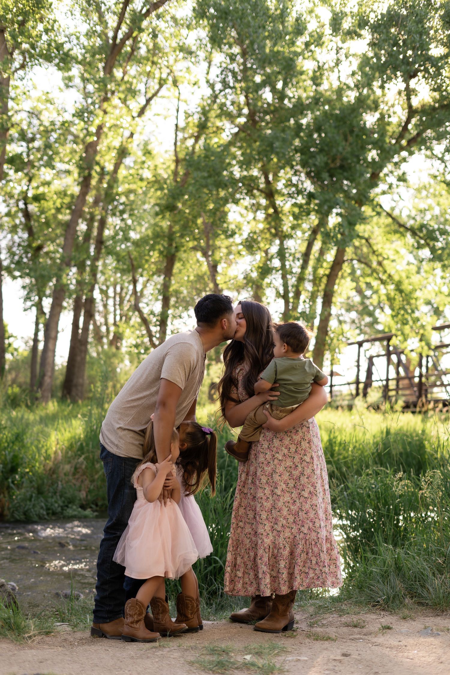 A young family shares a kiss outdoors, with a girl in a pink dress and cowboy boots and a toddler held by mom.