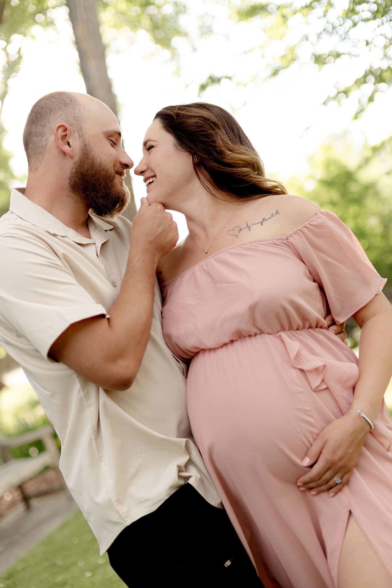 A smiling pregnant woman in a pink dress shares a tender moment with her bearded partner outdoors.