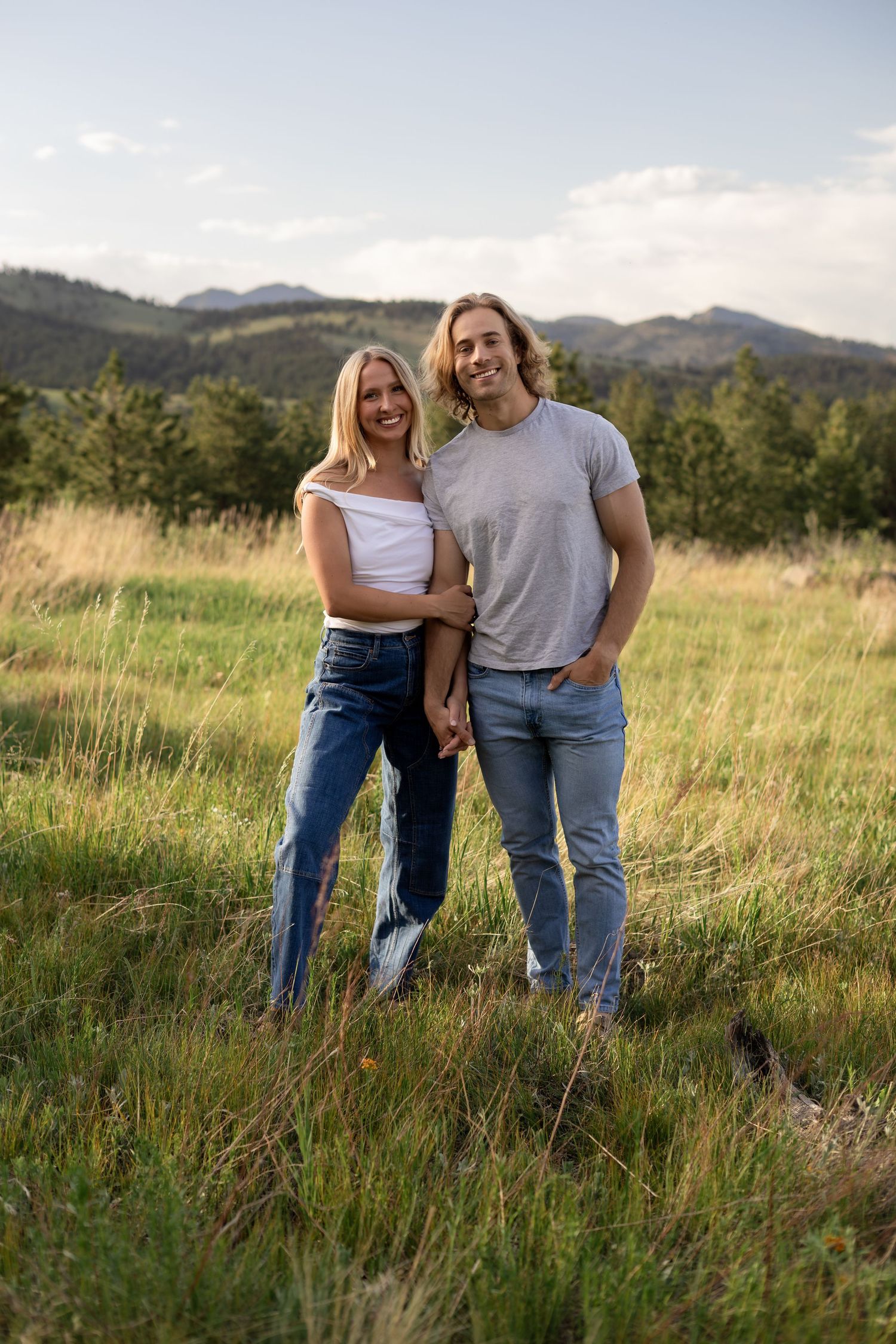 A smiling couple in jeans poses in a sunny mountain meadow, the woman in white top leaning into the man in gray tee.