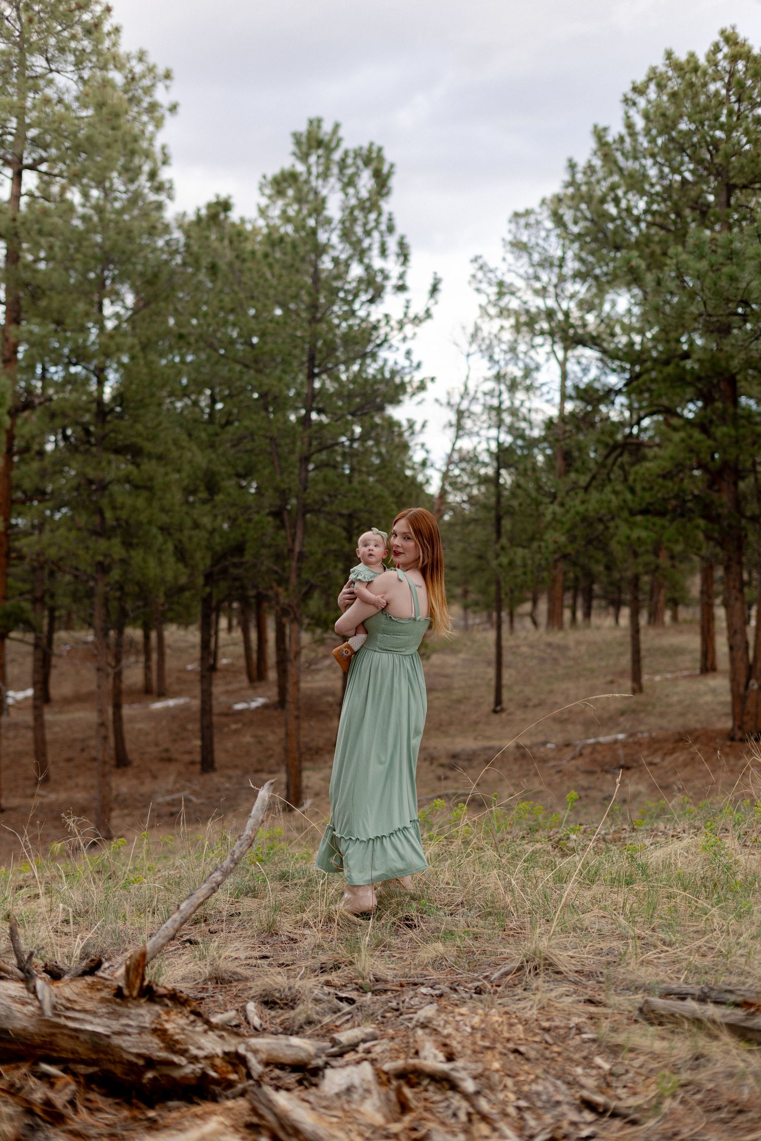 A woman in a sage green maxi dress holds a baby in a pine forest clearing under an overcast sky.