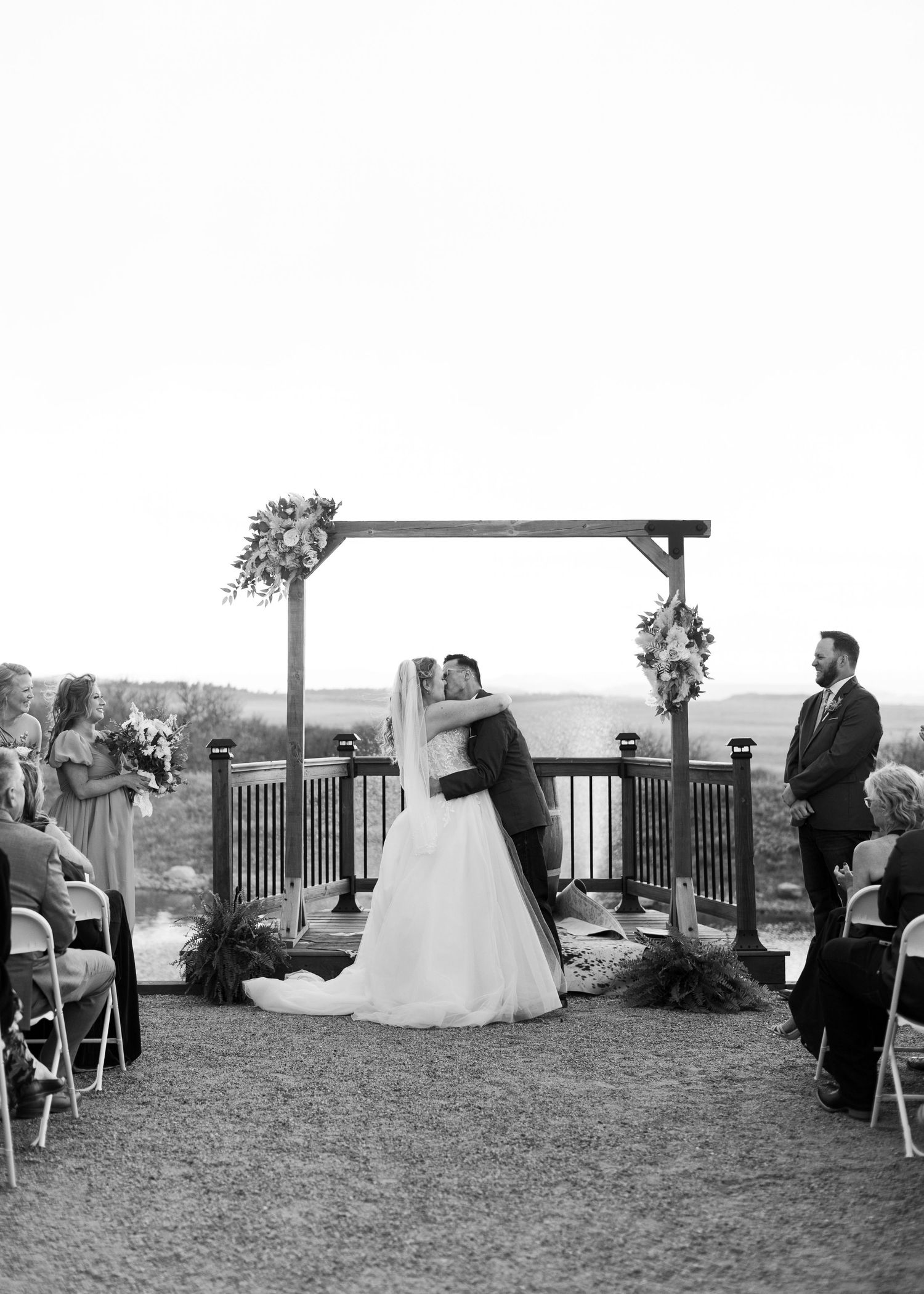 Black and white photo of a bride and groom sharing their first kiss at an outdoor wedding ceremony under a wooden arch.