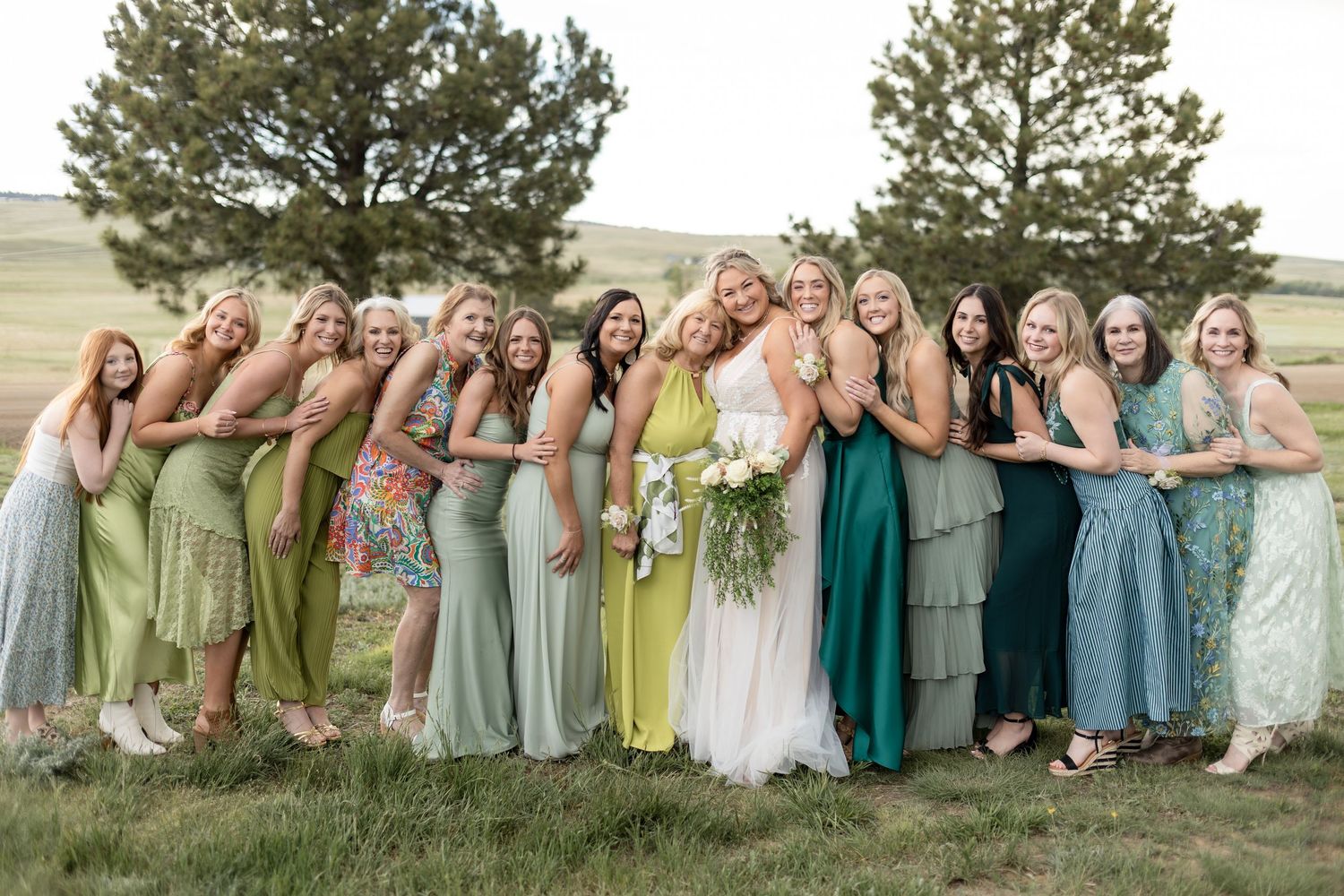 Bride poses with bridesmaids in green dresses outdoors on a grassy hill with pine trees in background.