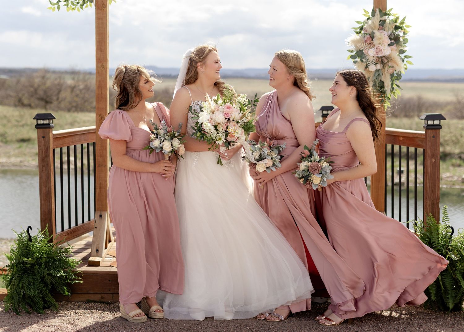 Bride in white gown with three bridesmaids in dusty rose dresses holding bouquets on a decorated wooden deck.