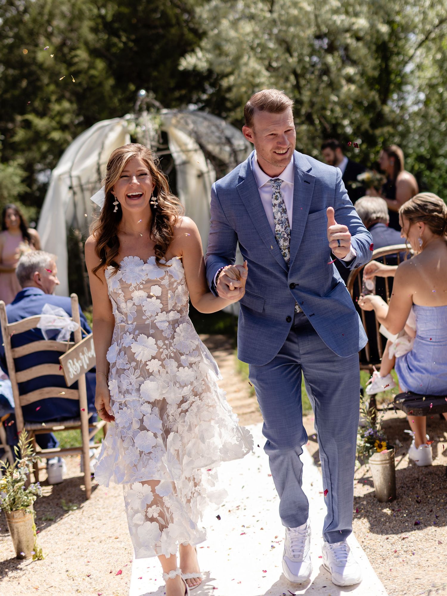 Joyful newlyweds walk down outdoor wedding aisle, bride in floral white dress, groom in blue suit, guests cheering.