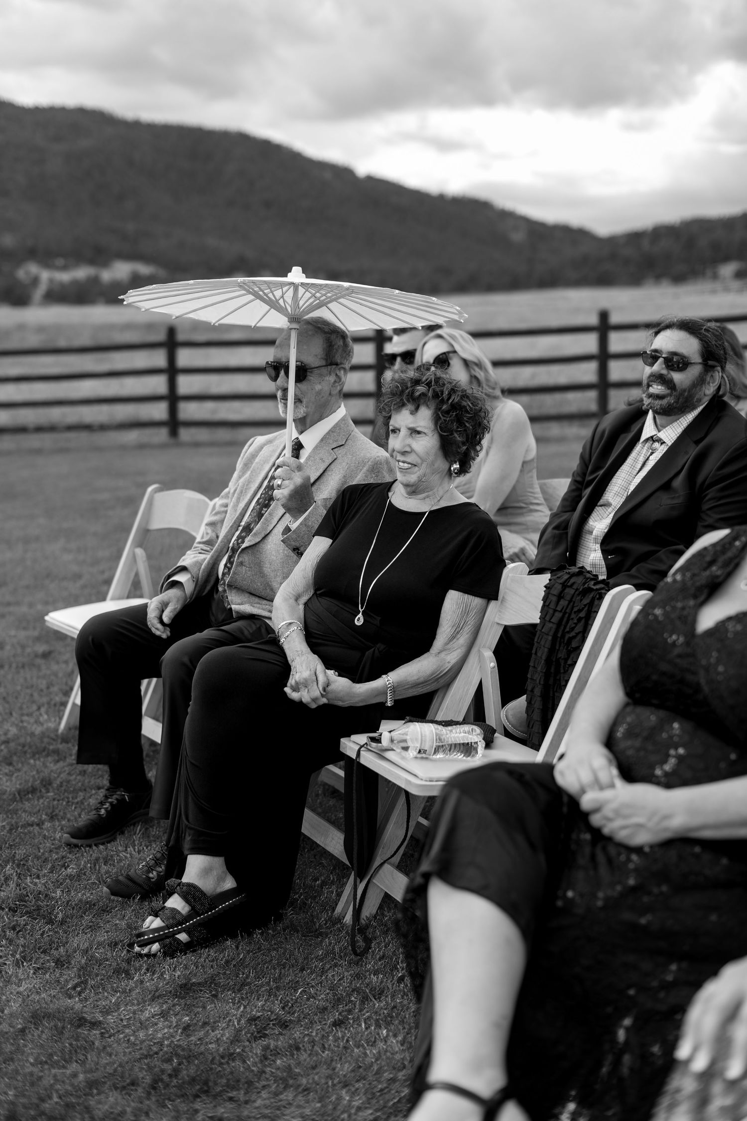 Wedding guests seated outdoors on white chairs, woman with parasol enjoying sunny ceremony.