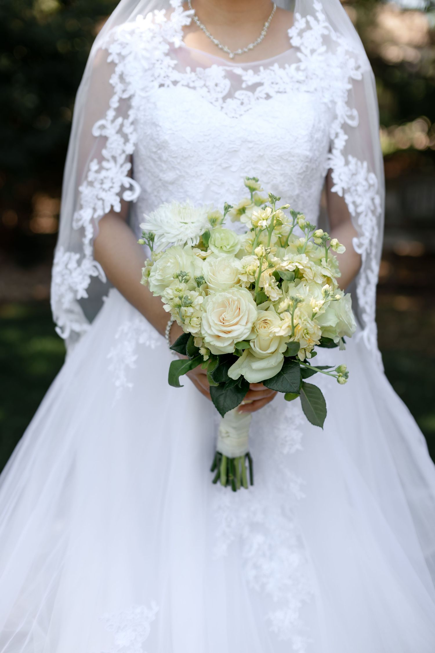 Bride in white lace wedding gown holding elegant bouquet of cream roses and green flowers.