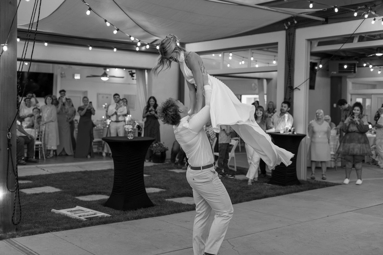 Groom lifting bride during first dance at wedding reception with guests watching in black and white photo.