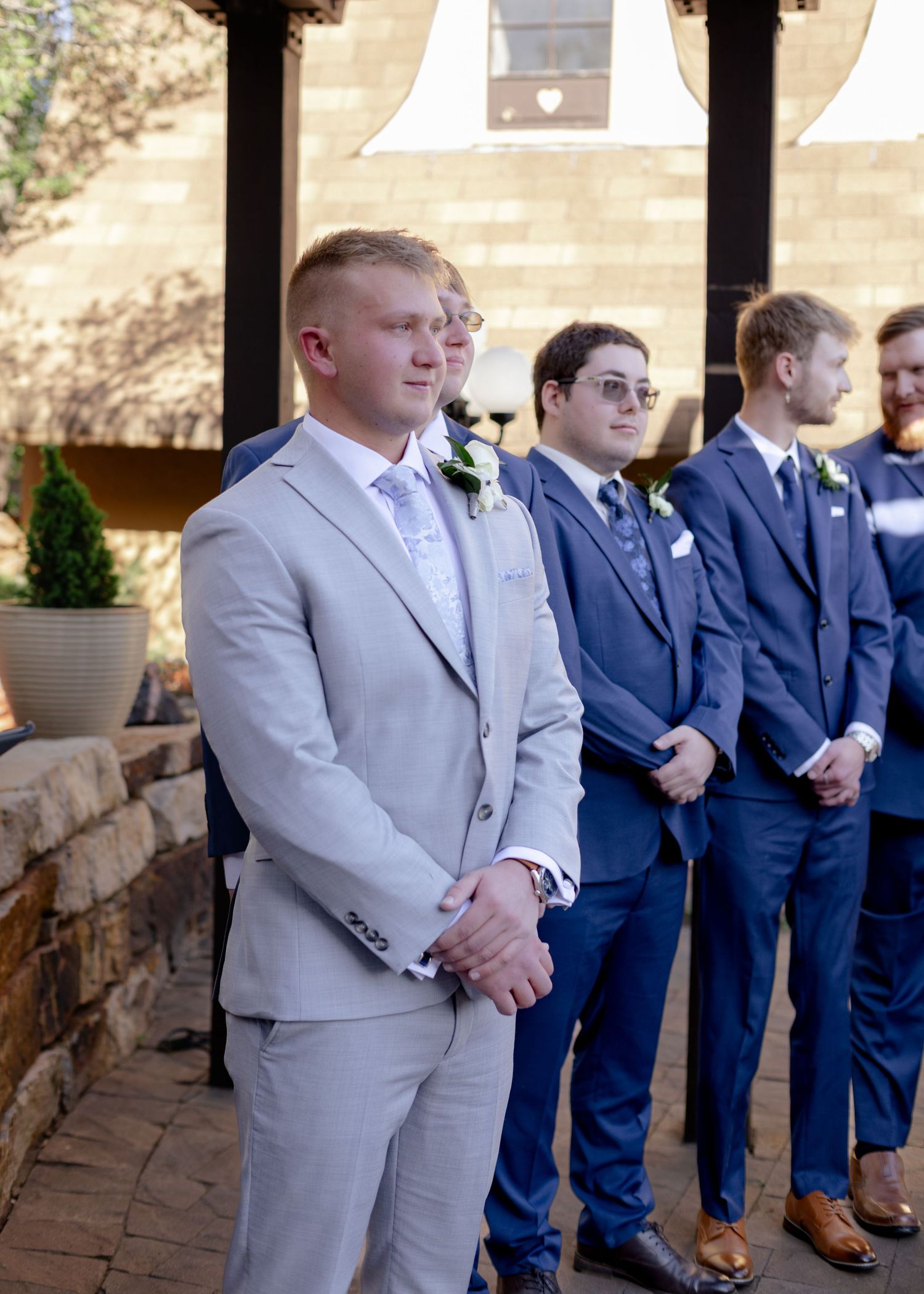 Groom in light gray suit stands with groomsmen in navy suits at an outdoor wedding ceremony venue.