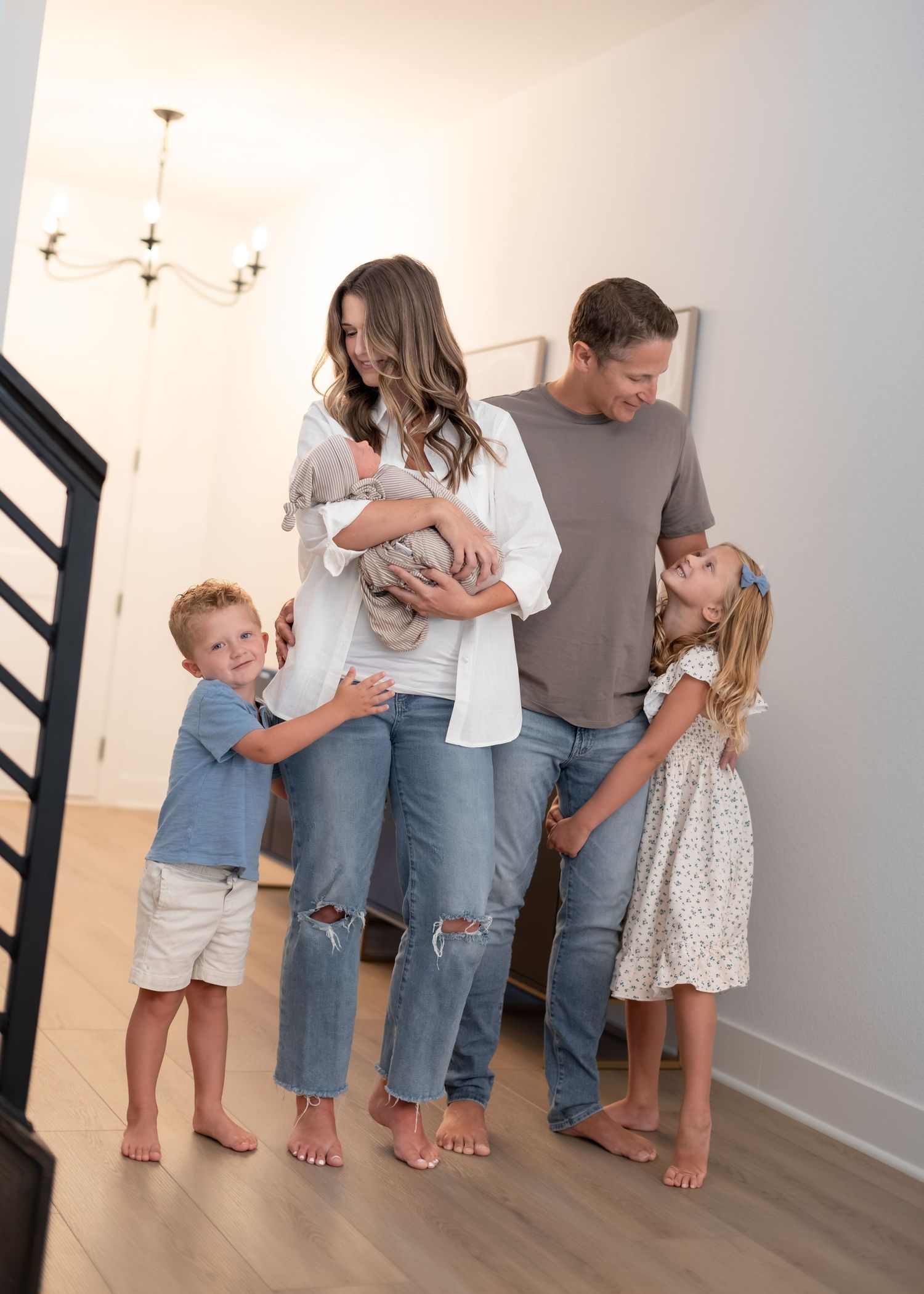 A happy family of five poses indoors, parents holding newborn baby while two young children smile and embrace them.