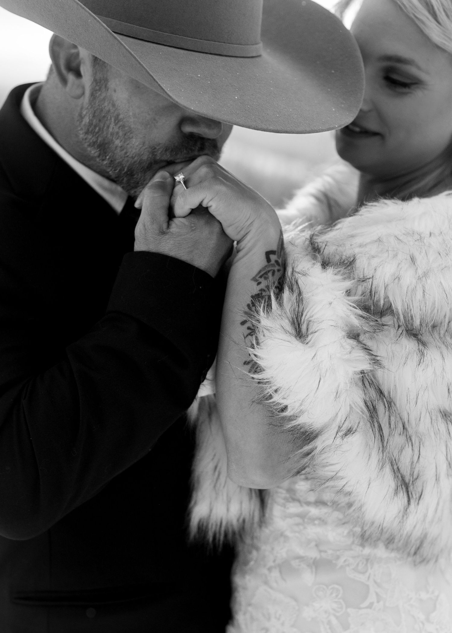 Black and white photo of a man in a wide-brim hat kissing a tattooed woman's hand, she wears a fluffy white coat.